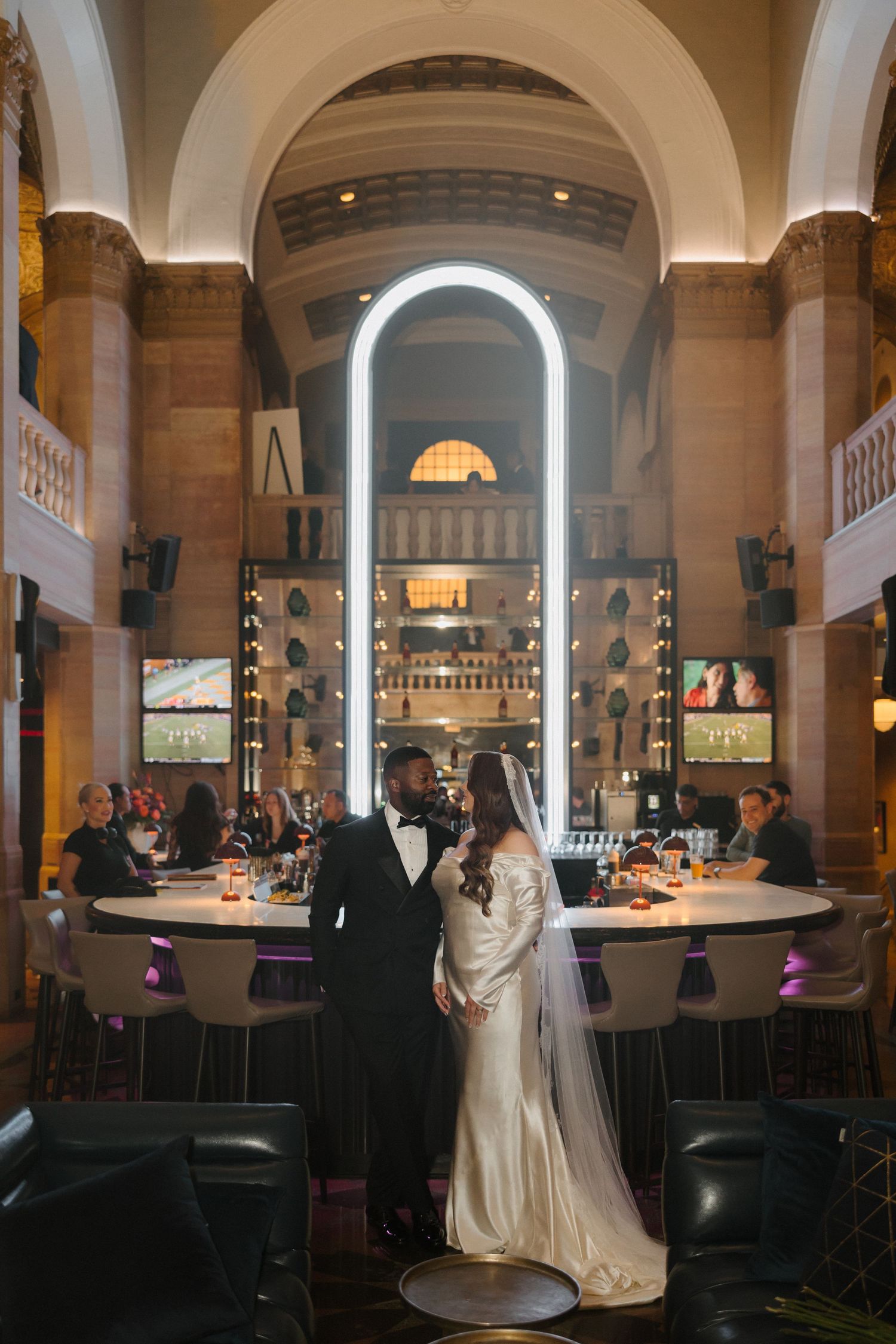 Elegant bride and groom posing inside a grand venue with arched ceilings, a glowing bar backdrop, and soft ambient lighting.