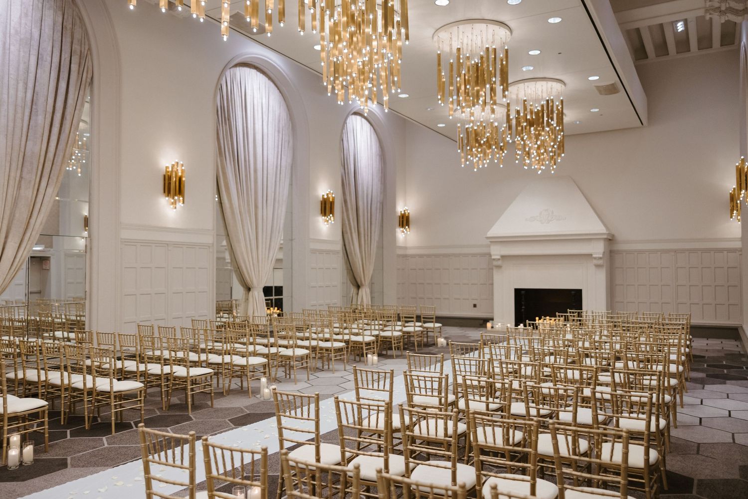 Elegant wedding venue ballroom with gold chiavari chairs, crystal chandeliers, draped curtains, and a white marble fireplace.