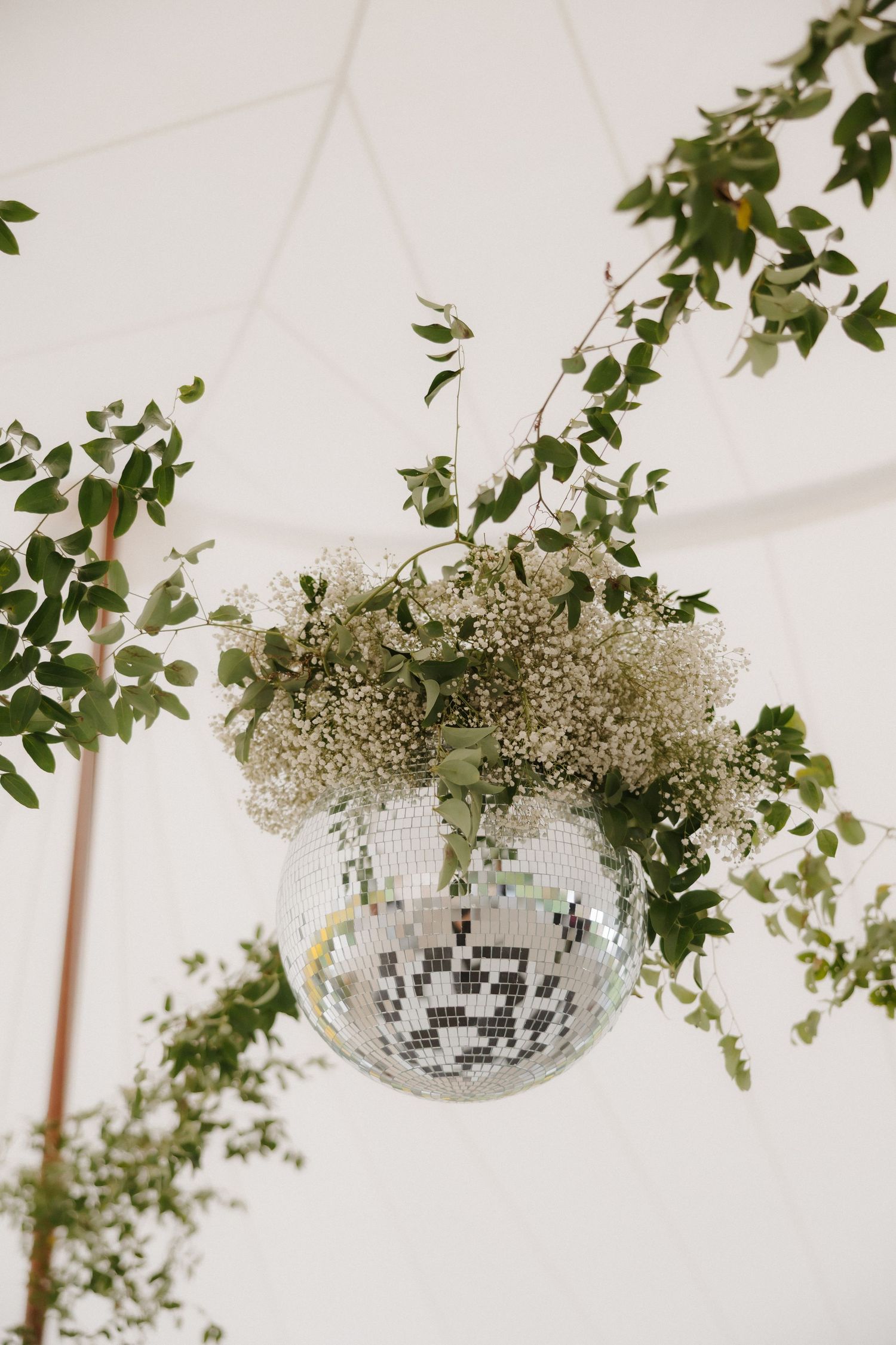 Close-up of a mirrored disco ball adorned with lush greenery and white blooms under a tent.