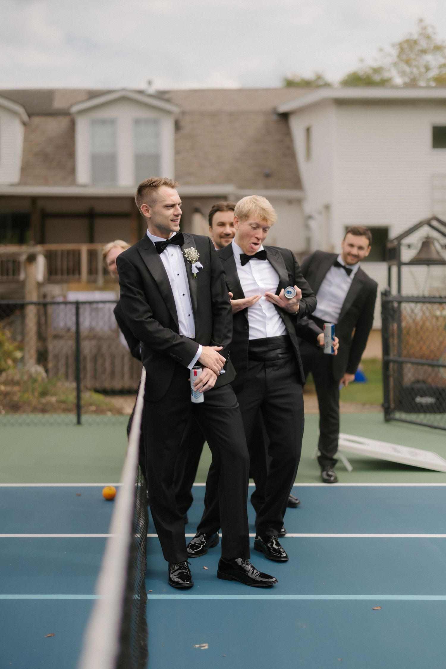 Groomsmen in black tuxedos playing pickleball on outdoor court before wedding ceremony.