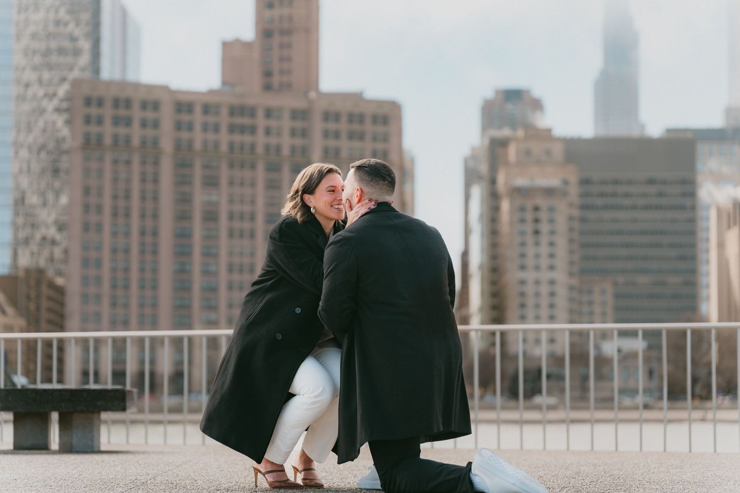 Romantic marriage proposal with man on one knee kissing his partner against Chicago city skyline backdrop.
