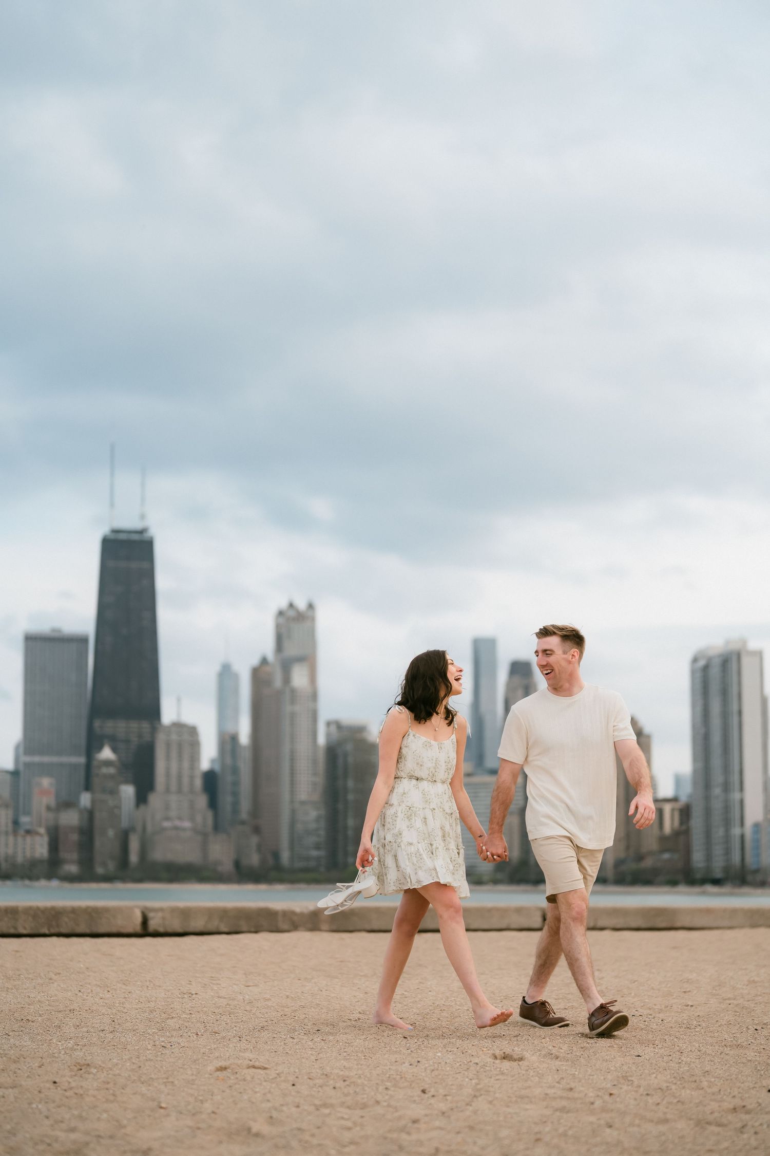 Couple holding hands and walking on a sandy beach with Chicago skyline backdrop.