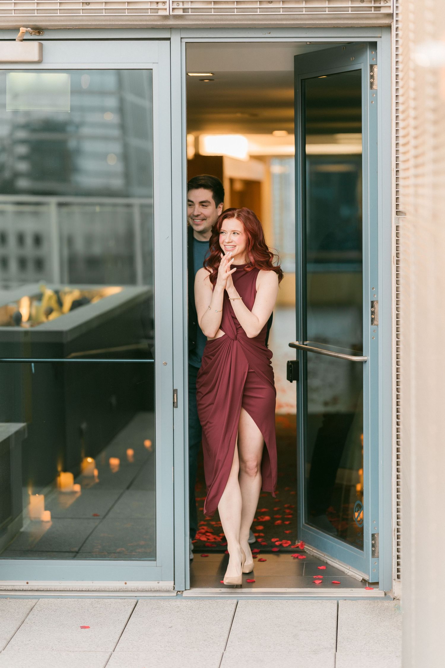 Smiling couple emerging through glass doors onto a rooftop with candles and rose petals at their feet.