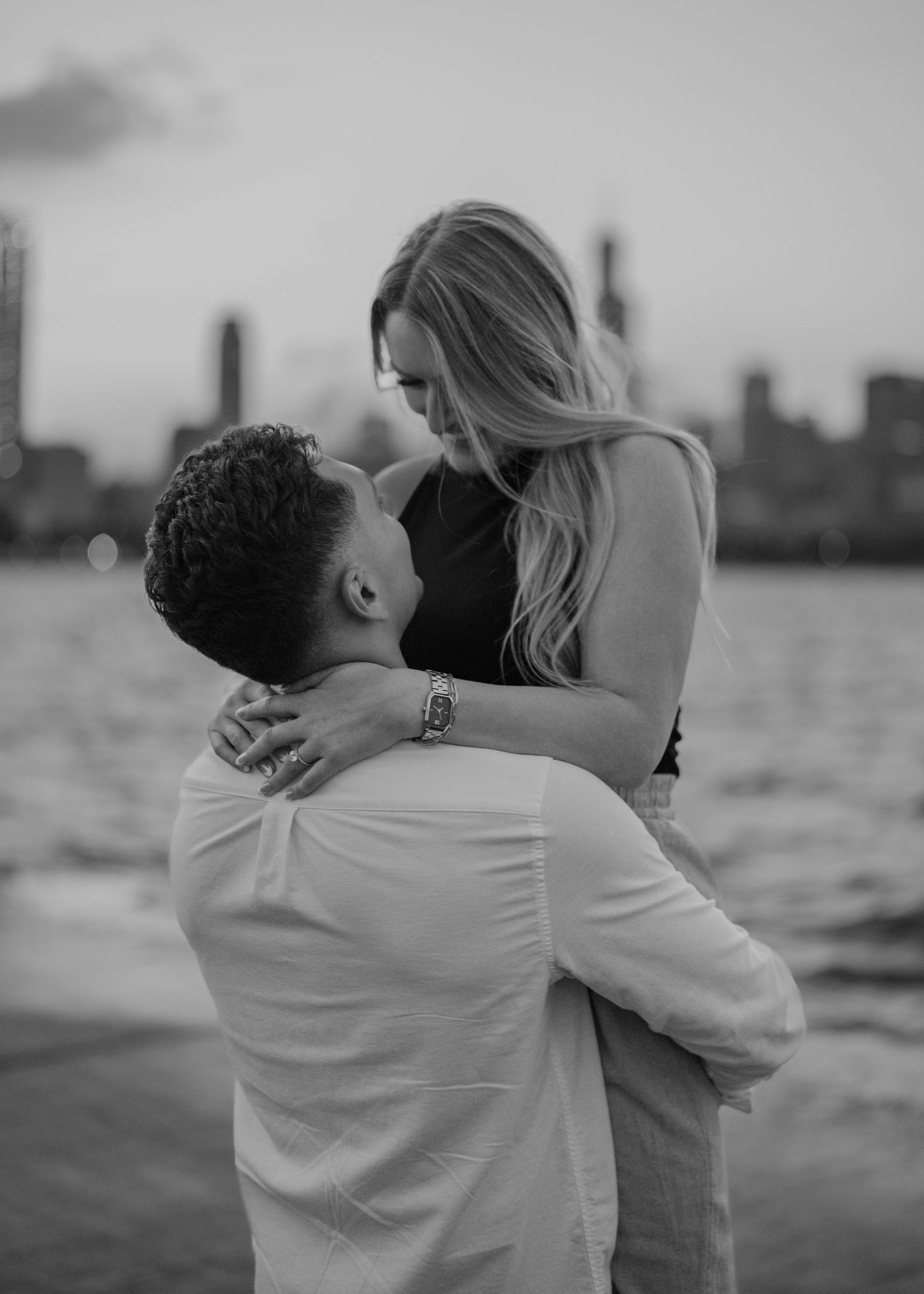 Black and white photo of a couple embracing near a waterfront with a city skyline at dusk.