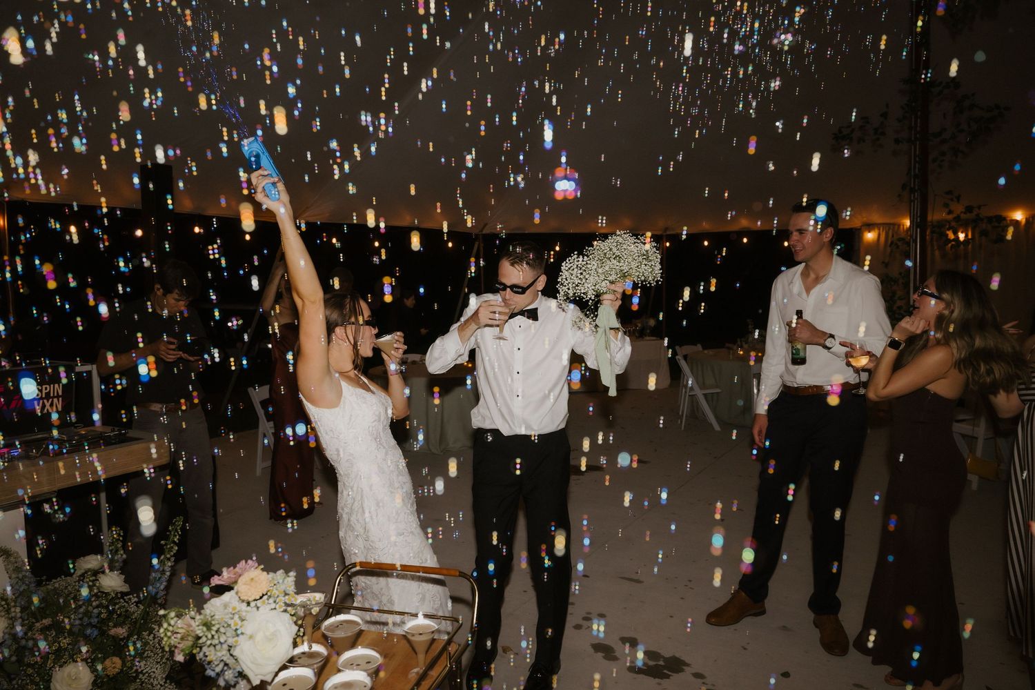 Bride and groom celebrate with confetti falling at lively wedding reception surrounded by guests holding drinks.
