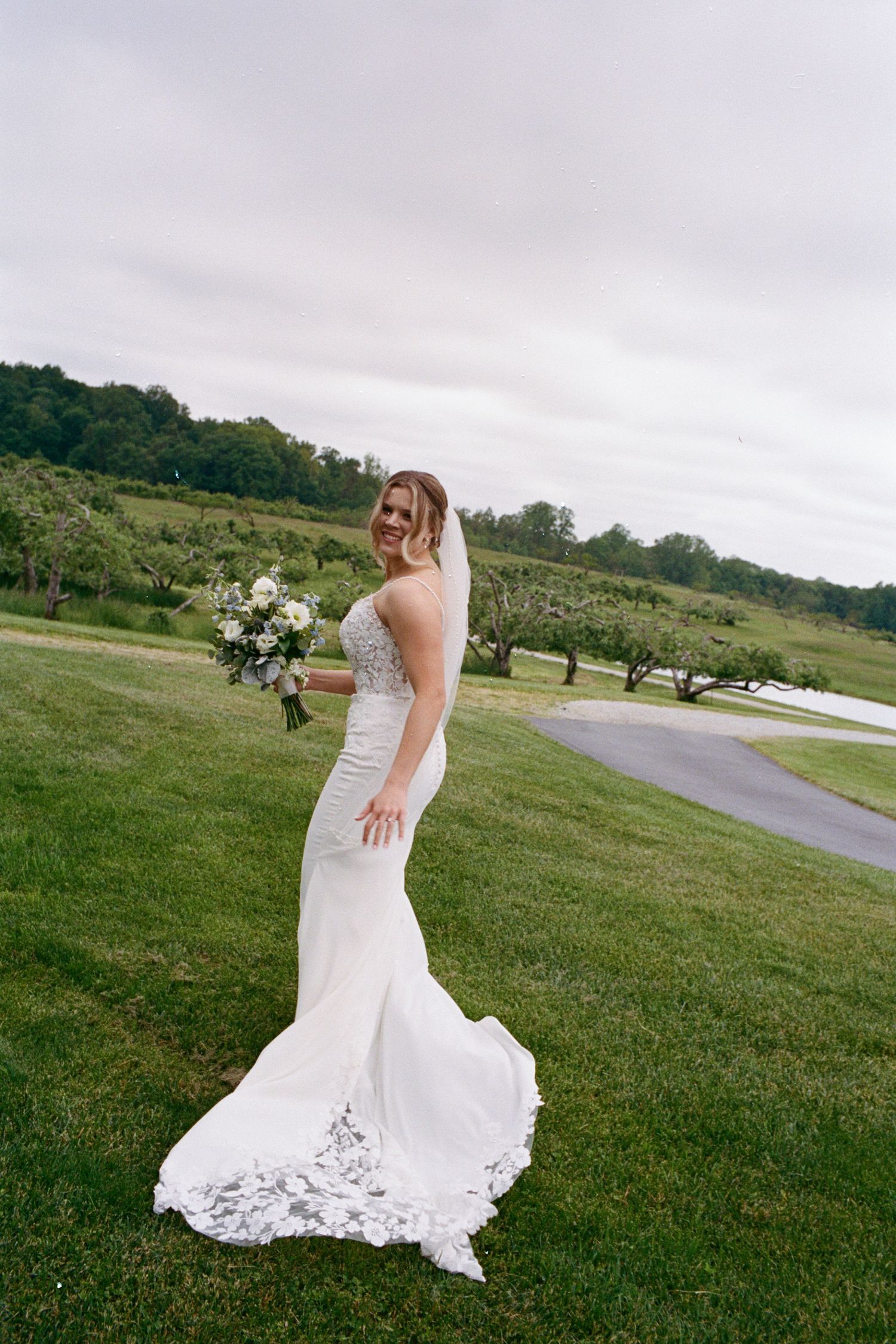 Bride in a white mermaid gown holding a bouquet, smiling outdoors on a green lawn with trees in the background.