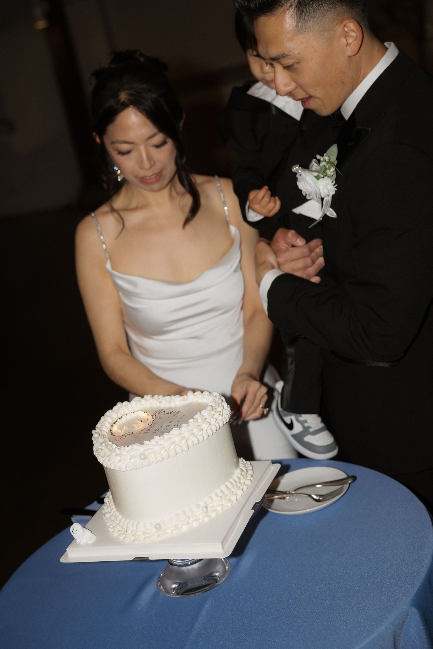 Asian bride and groom cutting white wedding cake together at reception, groom in tuxedo, bride in elegant white gown.