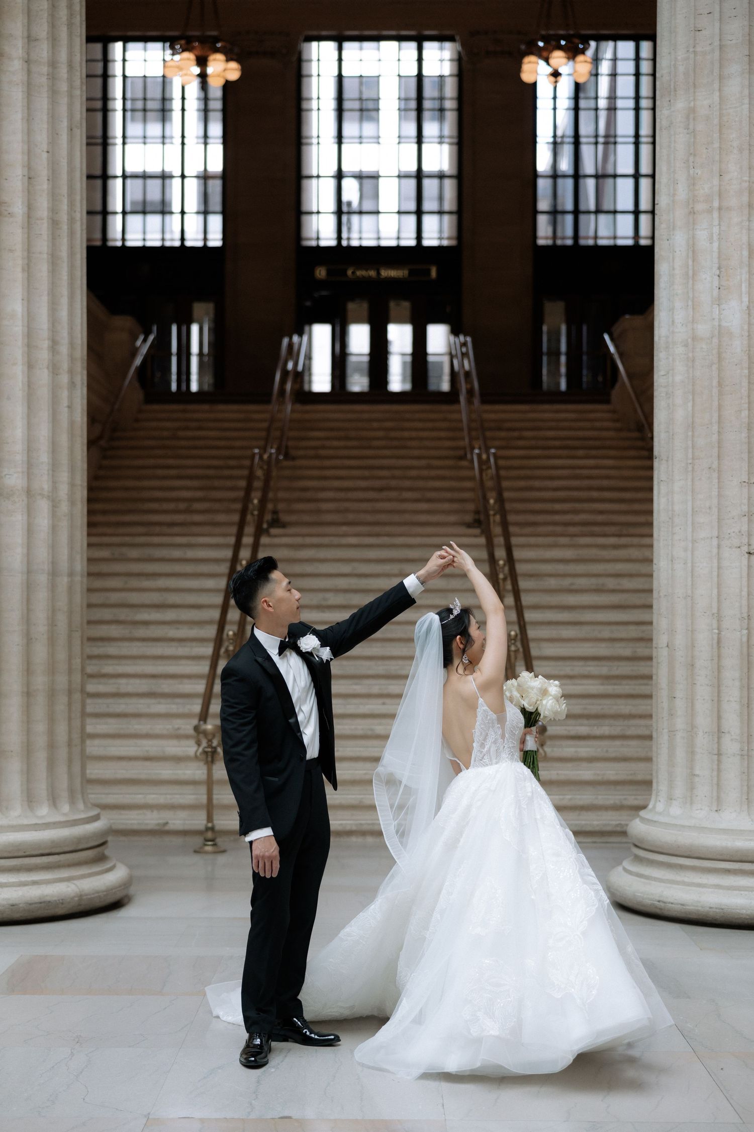 Groom twirls bride in stunning white gown inside grand marble hall with tall columns.
