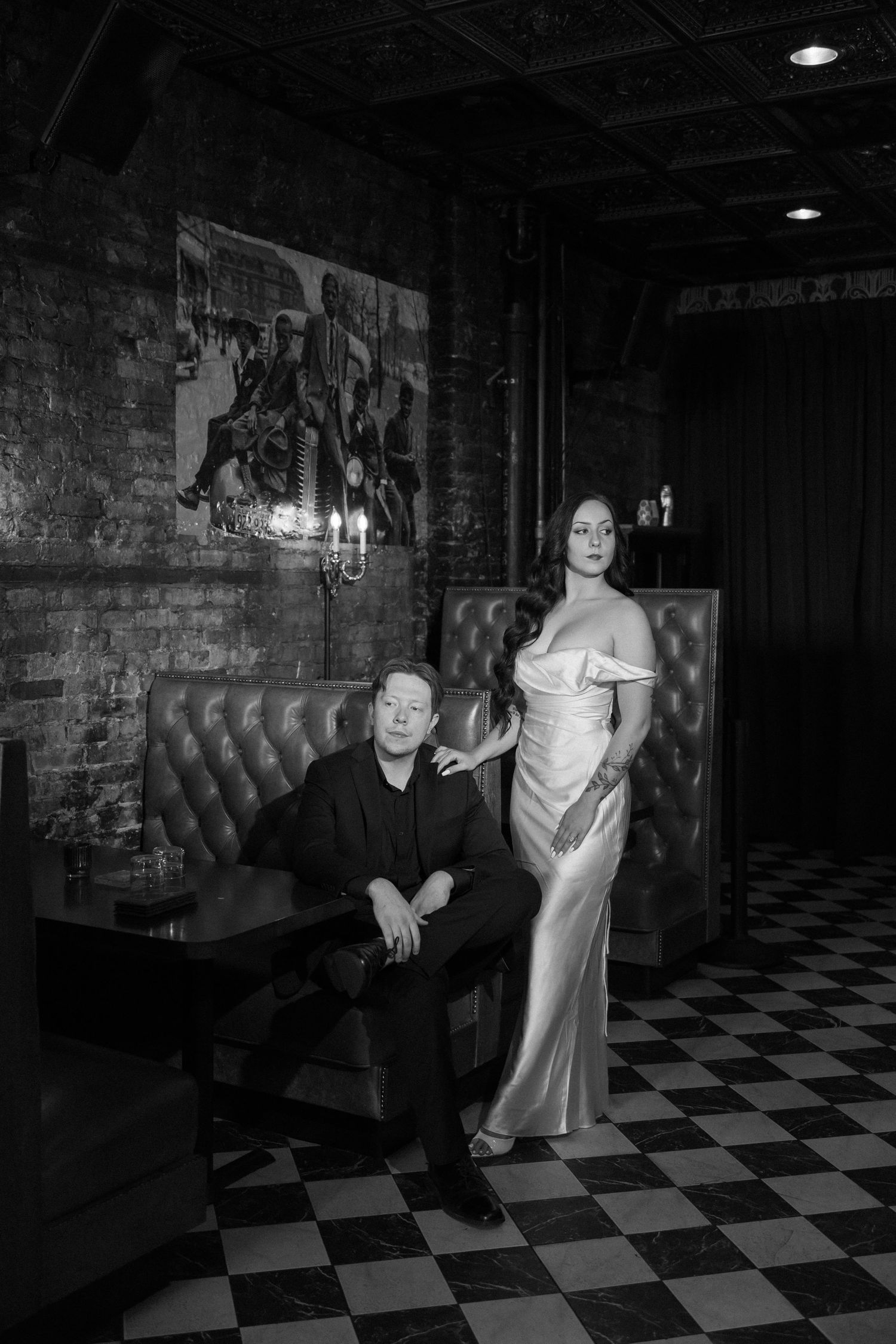 Black and white photo of elegant couple in upscale bar, man in suit seated, woman in satin gown standing beside him.