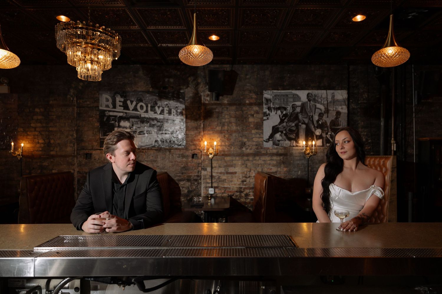 Man and woman sitting apart at a dimly lit bar with brick walls and vintage chandeliers.