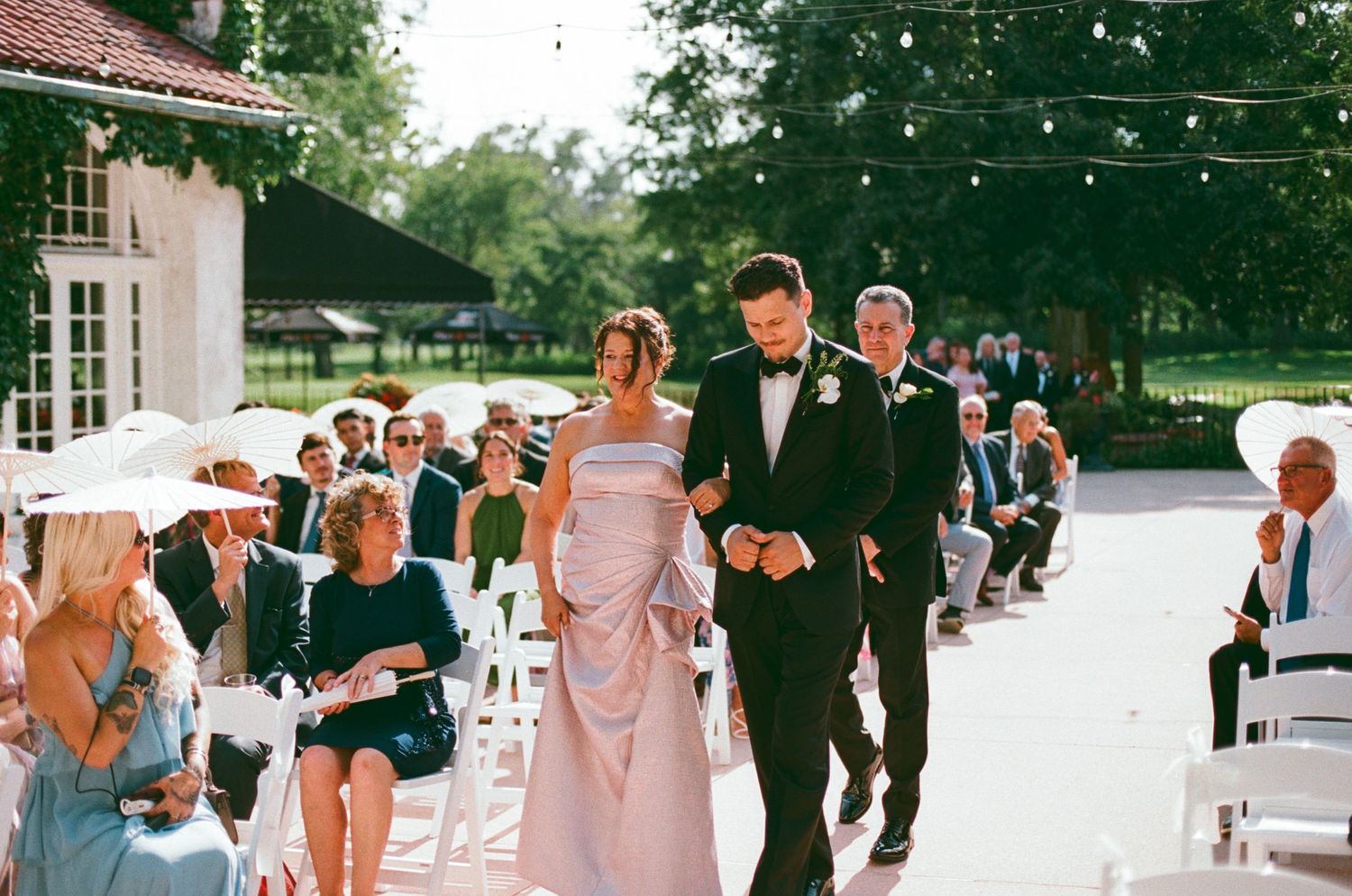 Bride in lavender gown walks down outdoor wedding aisle escorted by two men in tuxedos.