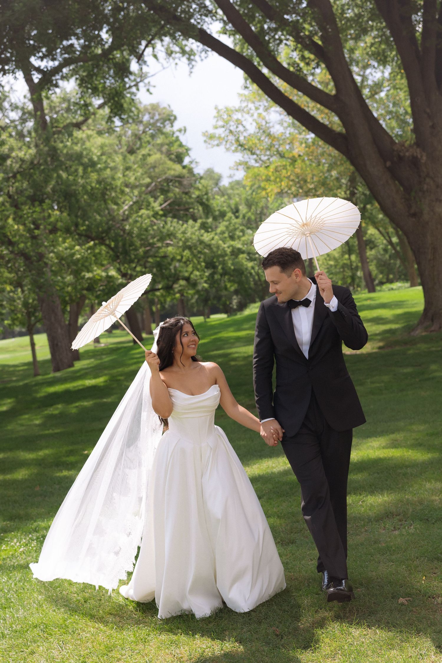 Bride and groom walking hand-in-hand in a lush green park, holding white parasols, she in a white gown, he in a black tuxedo.