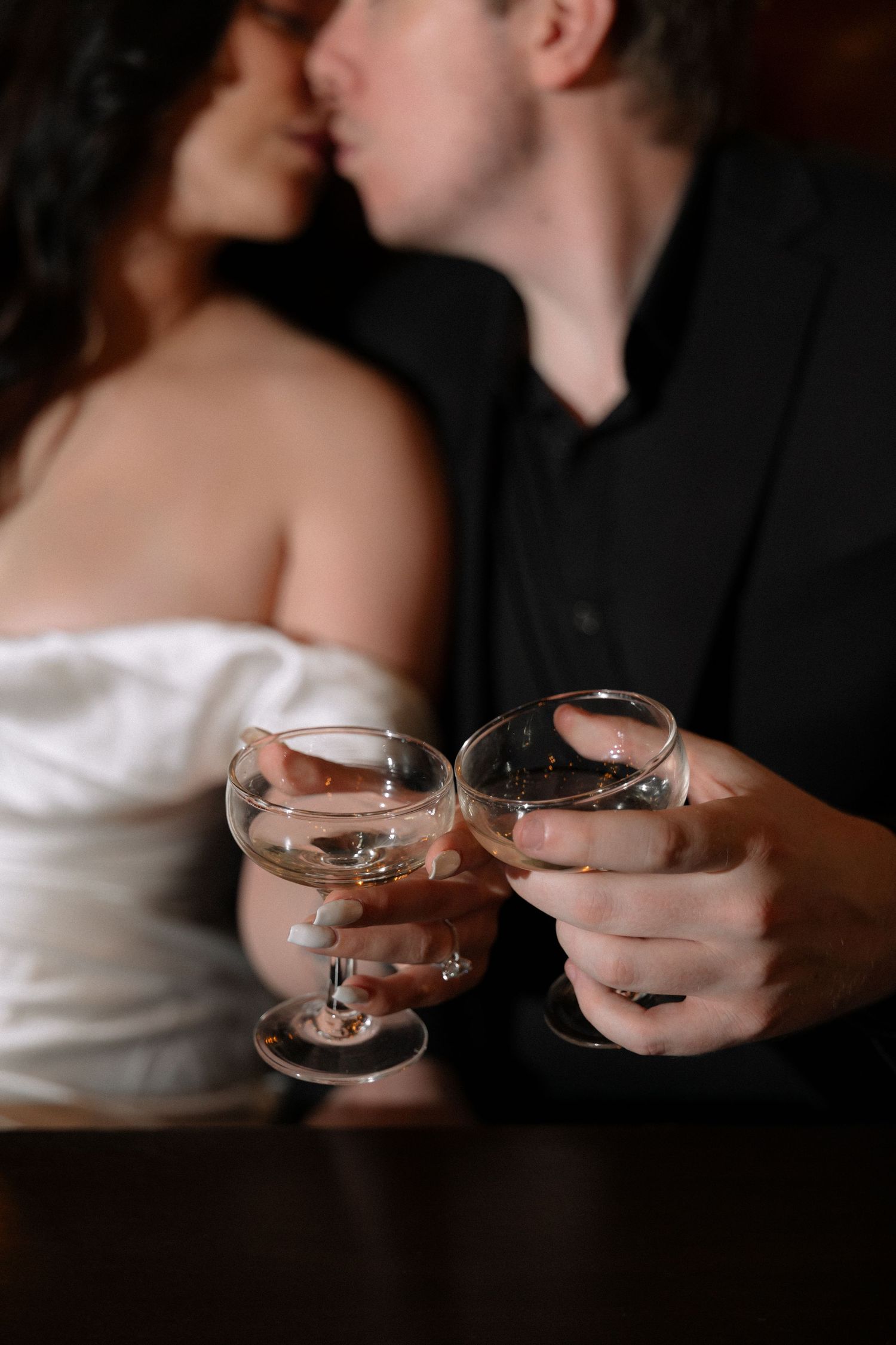 Close-up of a couple clinking champagne glasses while sharing a kiss in a dimly lit romantic setting.