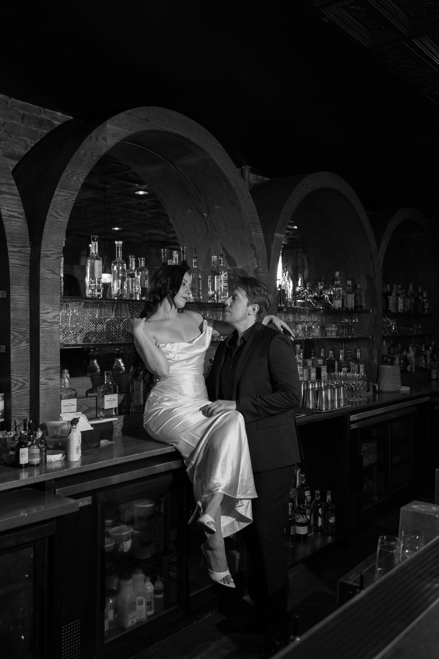 Black and white photo of bride sitting on bar counter in satin dress with groom standing close in elegant venue.