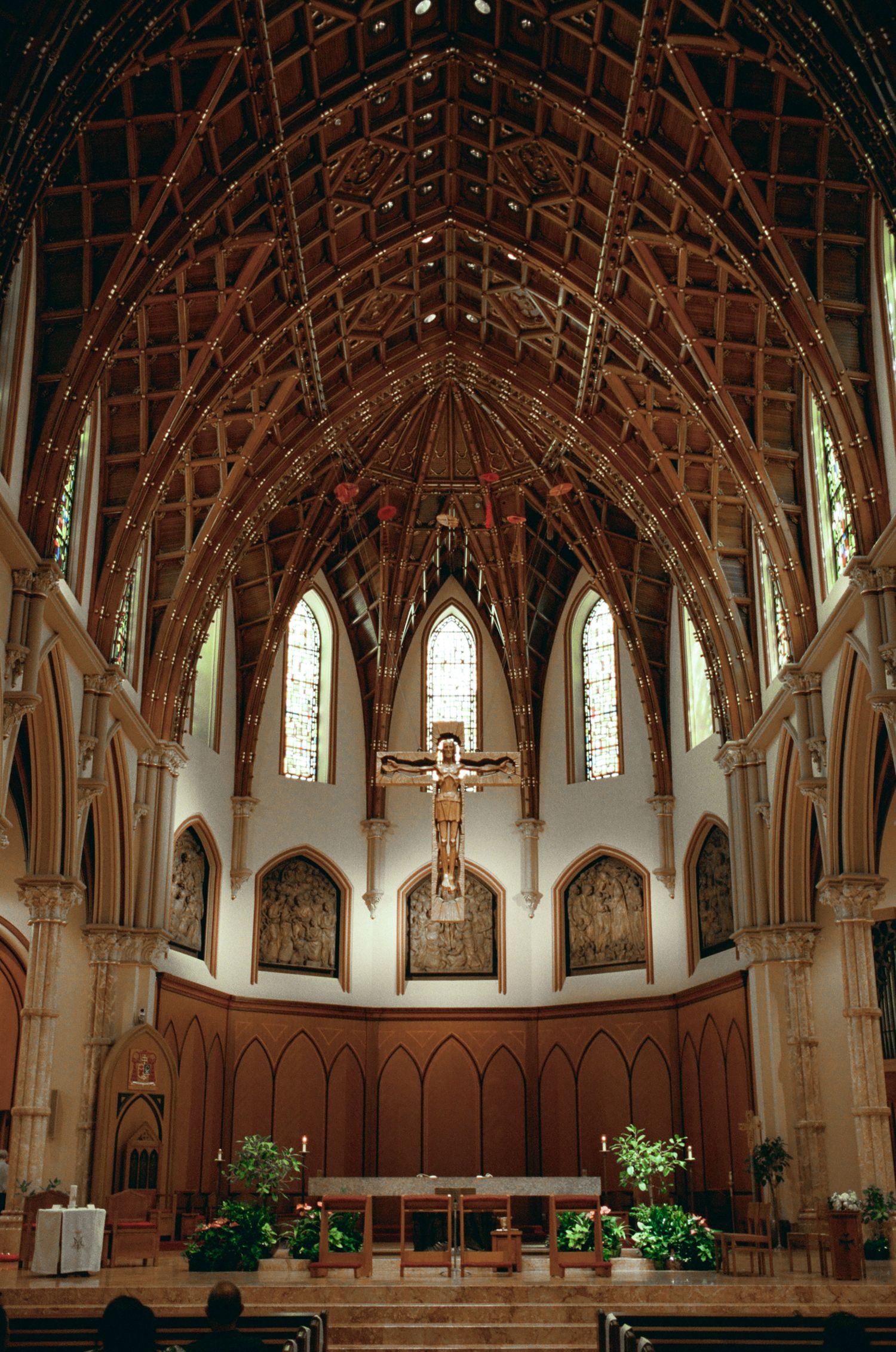 Gothic church interior with vaulted wooden ceiling, crucifix, stained glass windows, and floral altar decorations.