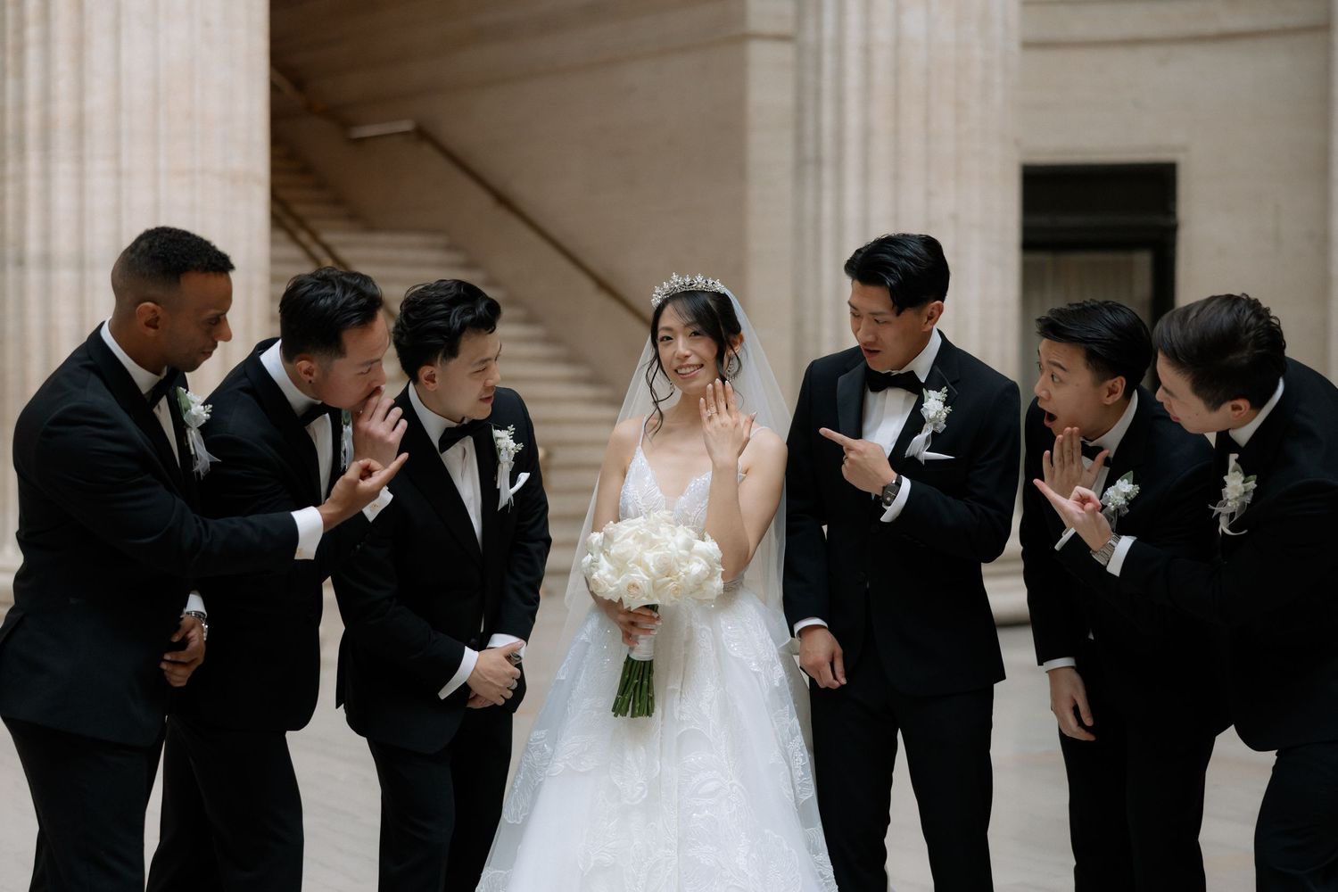 Asian bride in white gown with groomsmen in black tuxedos posing on marble steps outdoors.