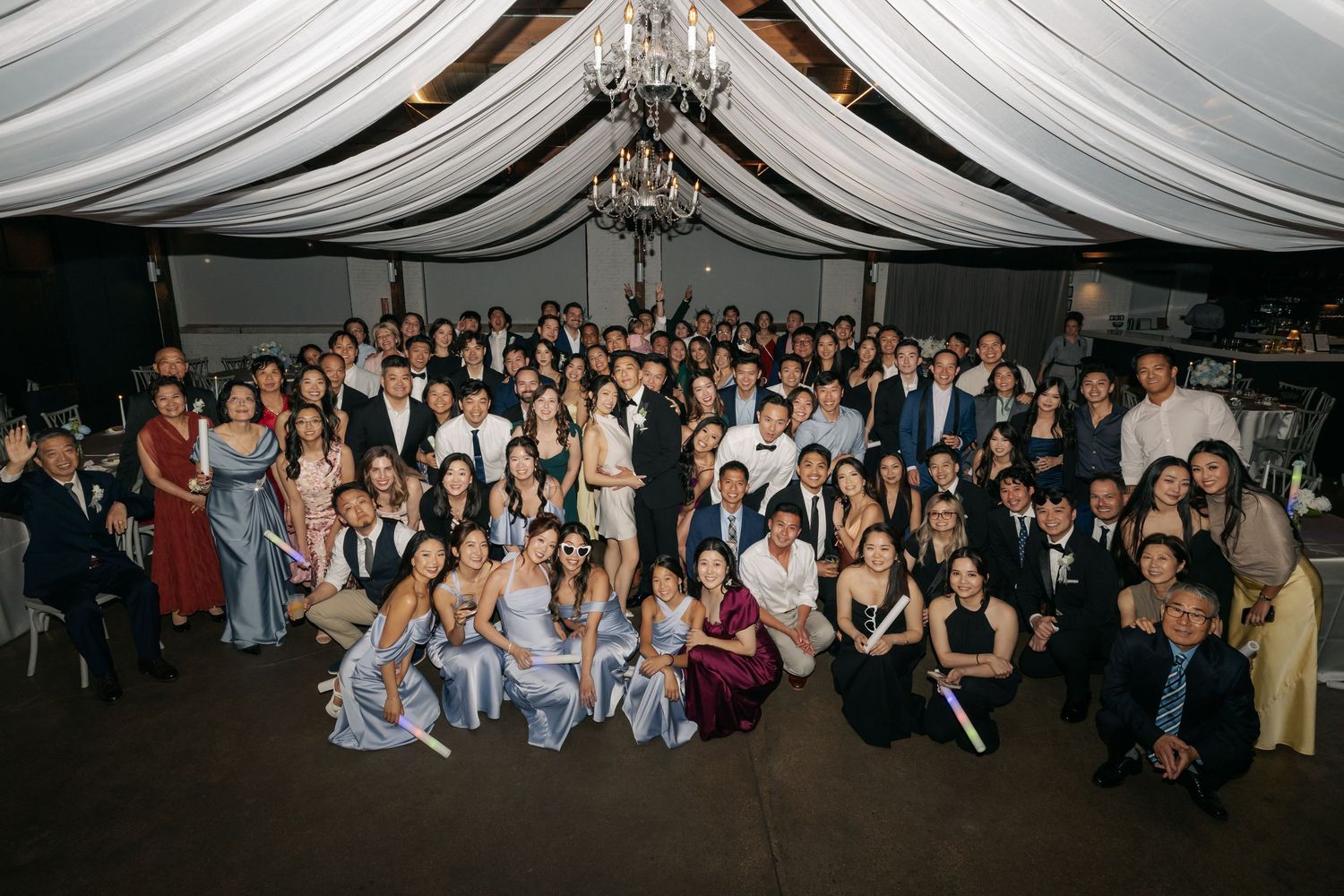 Large group wedding photo of guests gathered under draped ceiling at reception venue.