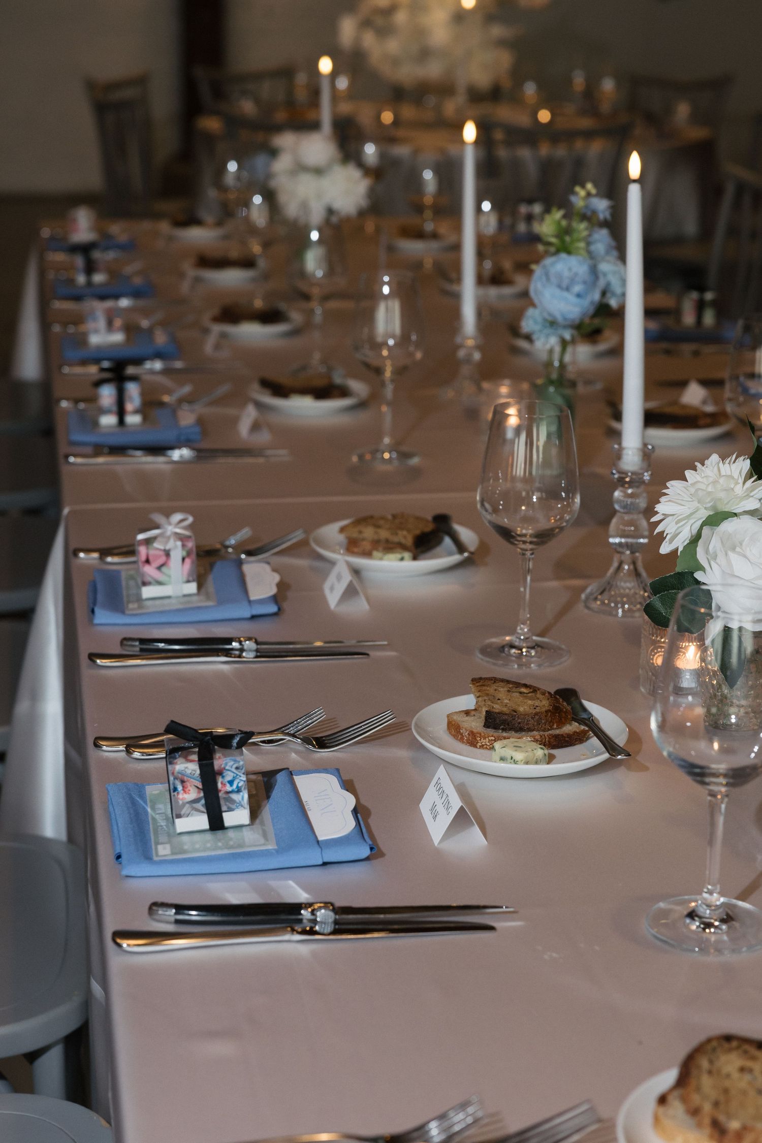 Elegant wedding reception table set with blue napkins, white candles, floral centerpieces, crystal glasses, and bread plates.