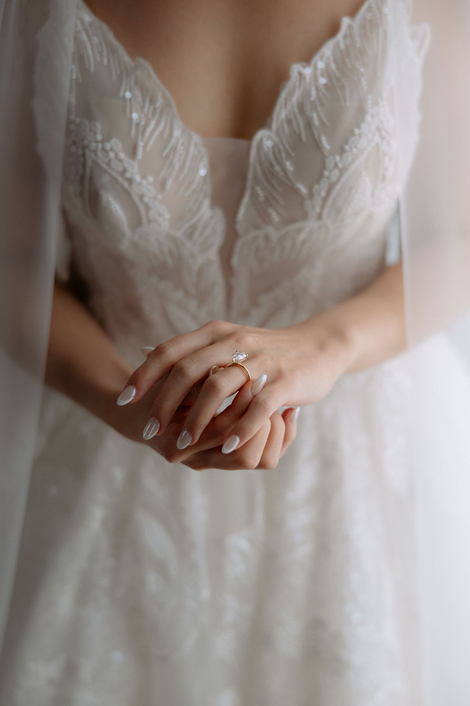 Bride in lace wedding gown clasps hands showing diamond engagement ring with white manicure.