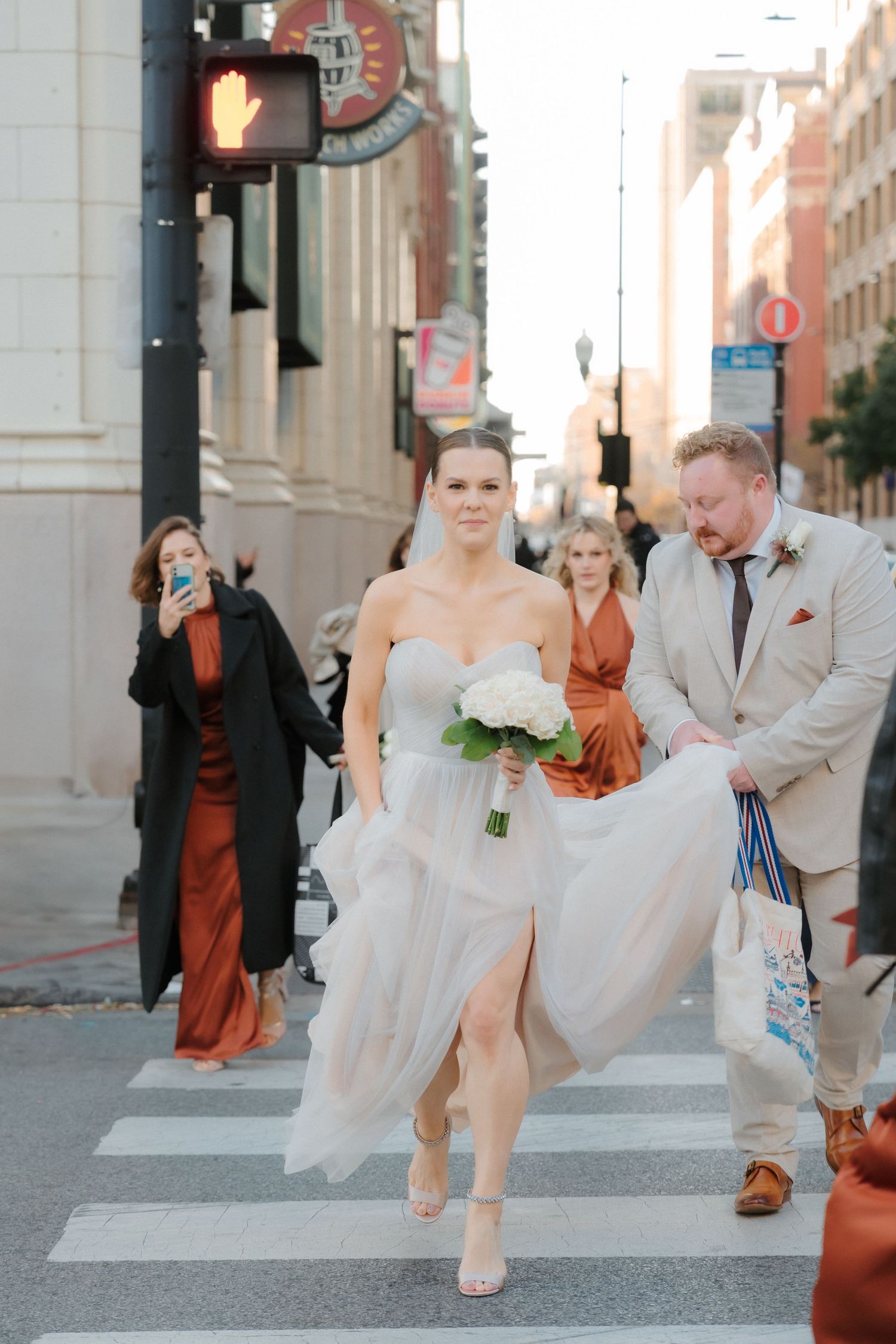 Bride in strapless gown crosses city street holding white bouquet with wedding party.