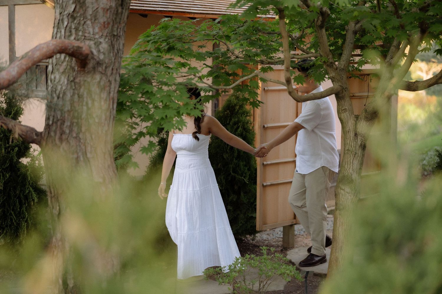 Couple in white attire sharing a romantic moment among lush green trees in a garden setting.
