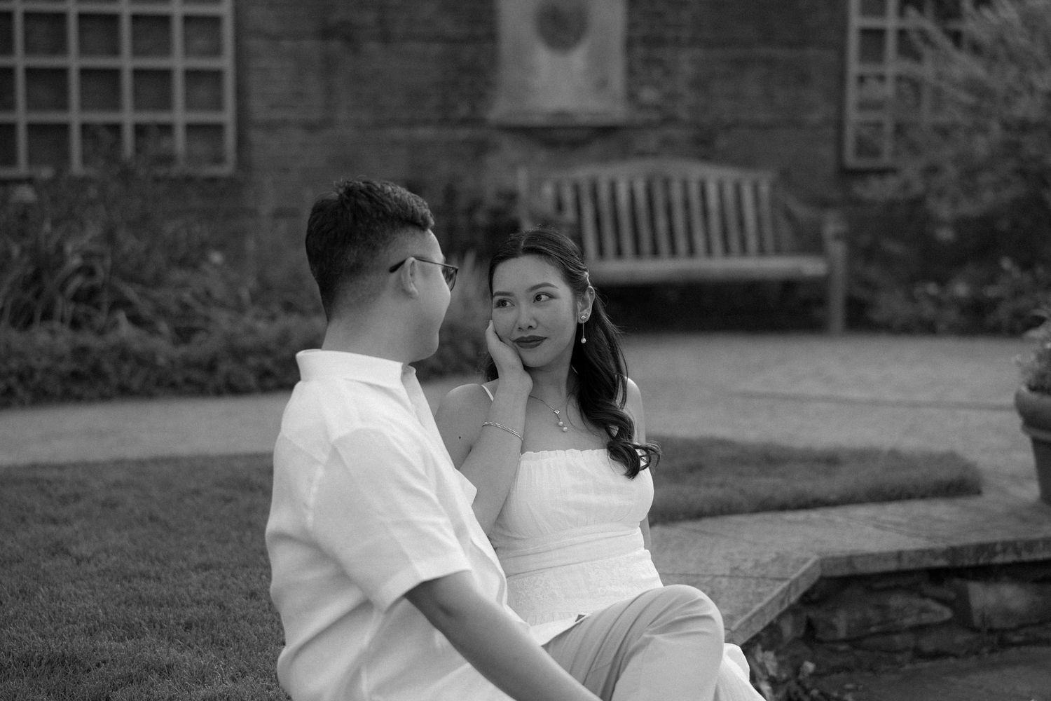 Black and white photo of a young couple sitting outdoors in a garden, sharing an intimate moment near a brick building.