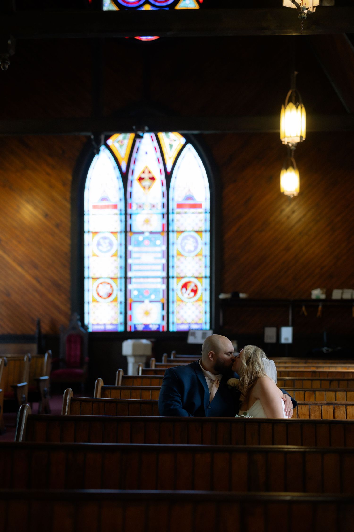 Couple sharing a kiss in wooden church pews with colorful stained glass window backdrop during wedding ceremony.