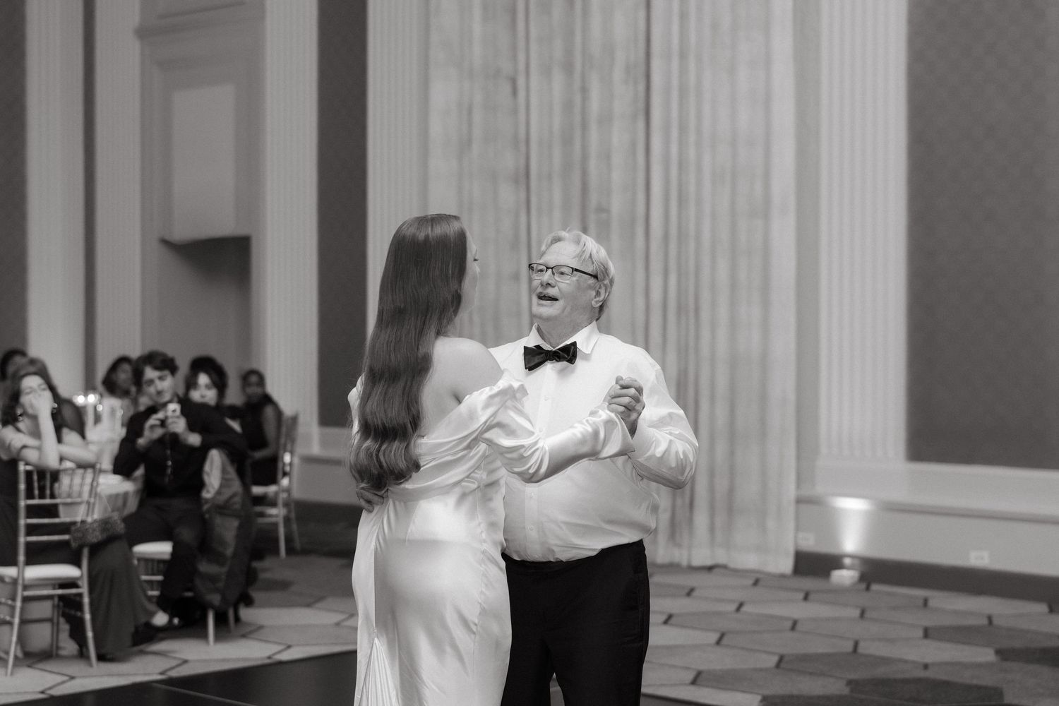 Father-daughter wedding dance in elegant ballroom, black and white photo of bride in white gown with older man in tuxedo.