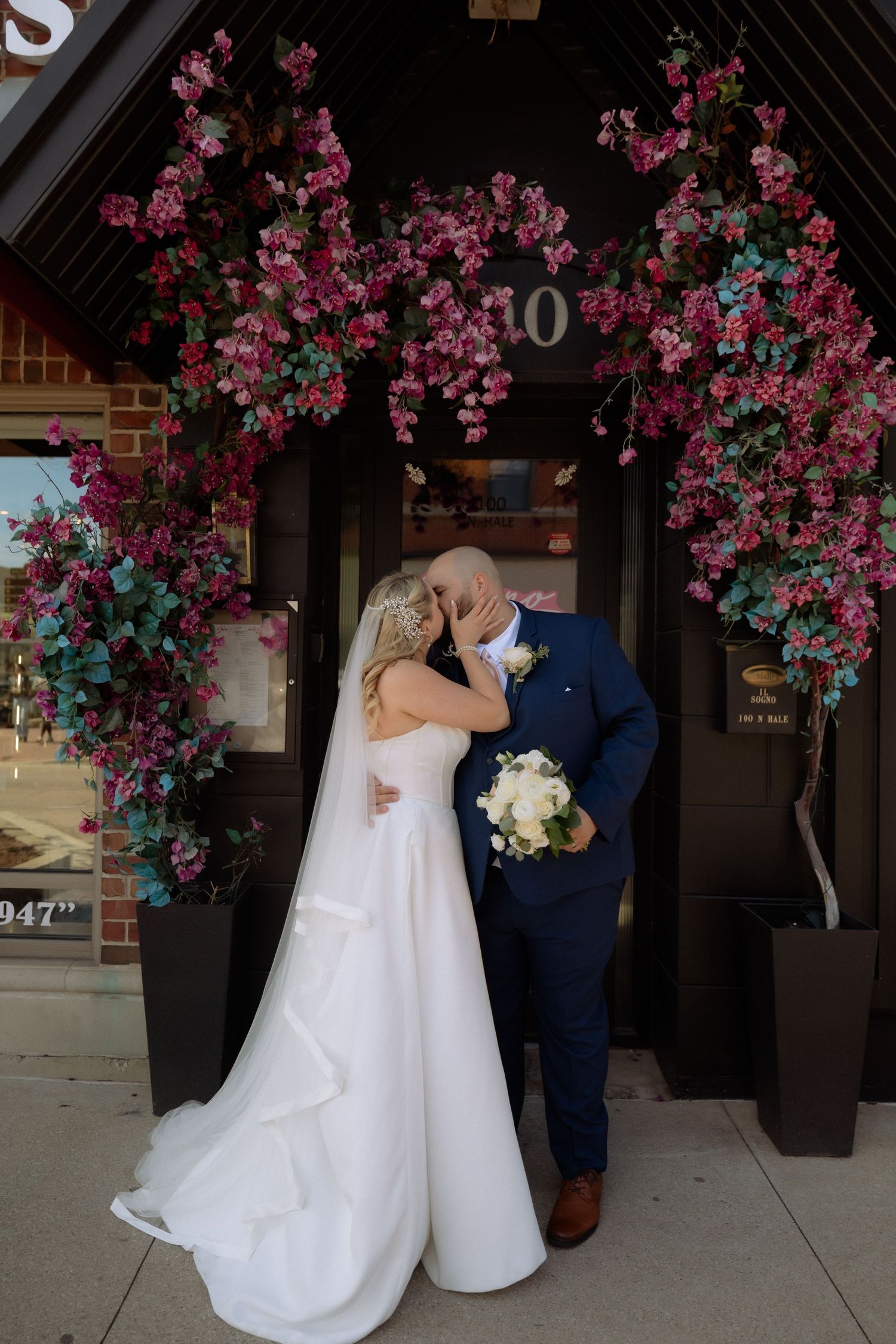 Bride and groom kissing under a floral arch with pink blooms at a brick building entrance on their wedding day.