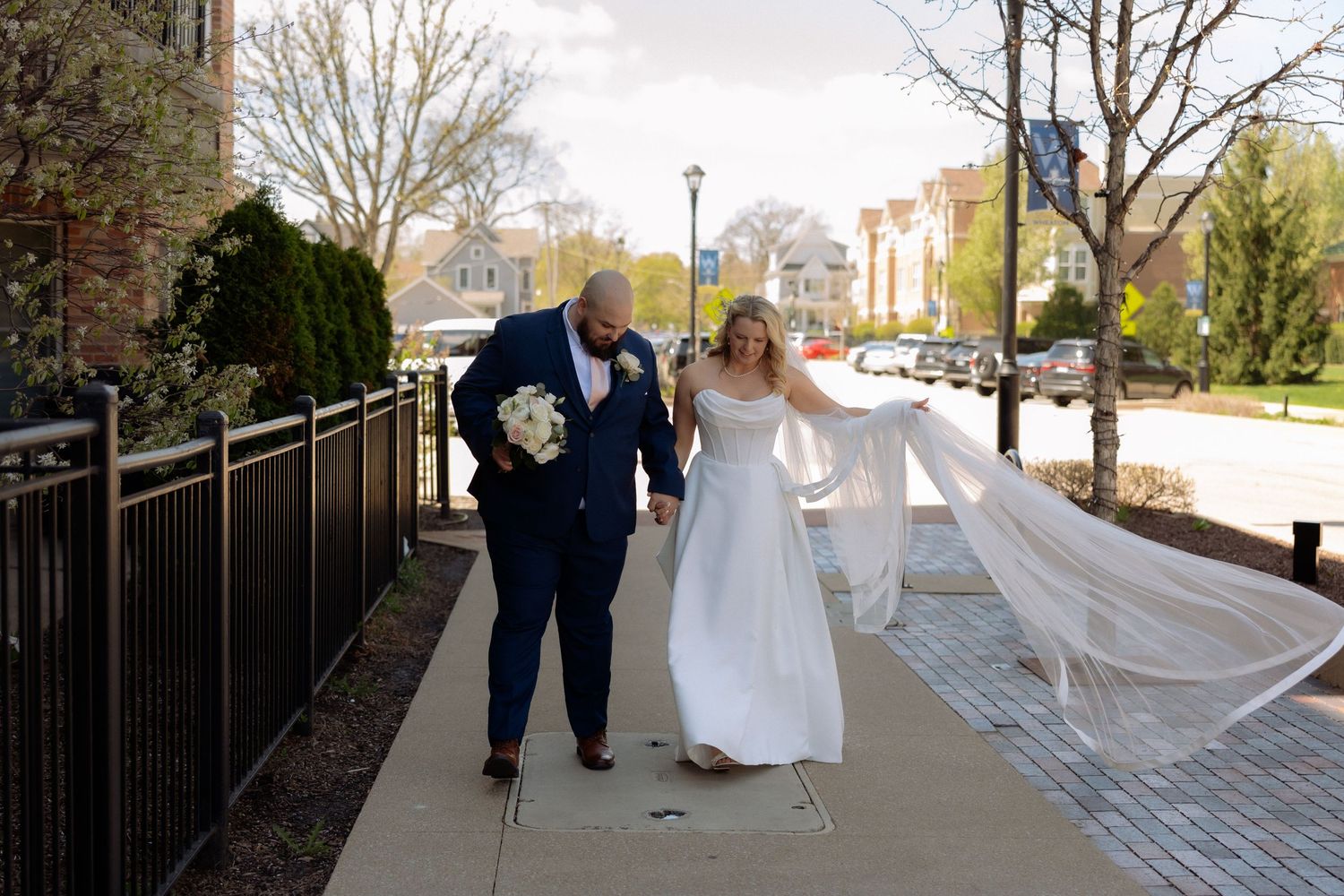 Bride and groom walking together outdoors on their wedding day, bride's veil flowing in the wind.
