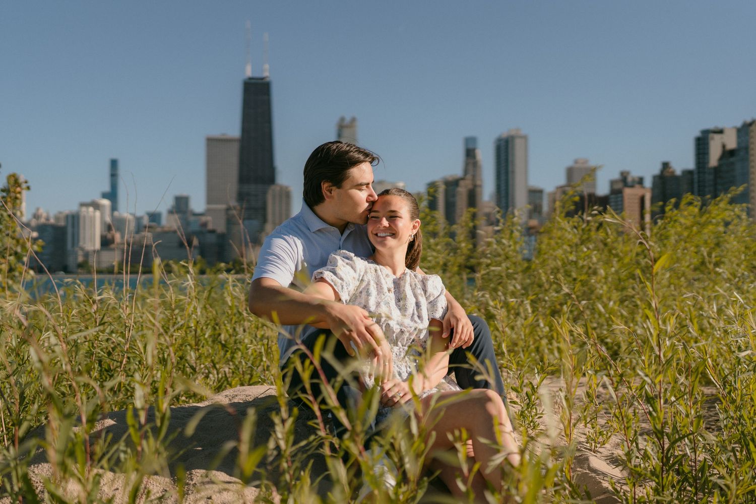 Couple sitting together among tall green grass with Chicago city skyline visible in the sunny background.