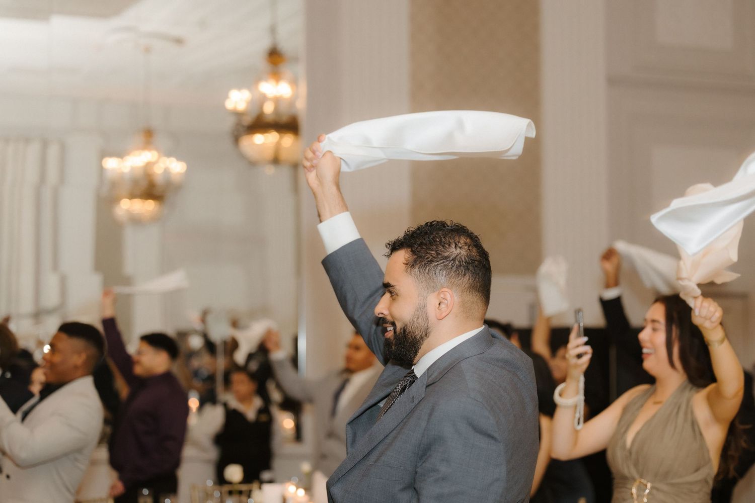 Wedding guests waving white napkins in elegant ballroom celebration during lively reception dance.