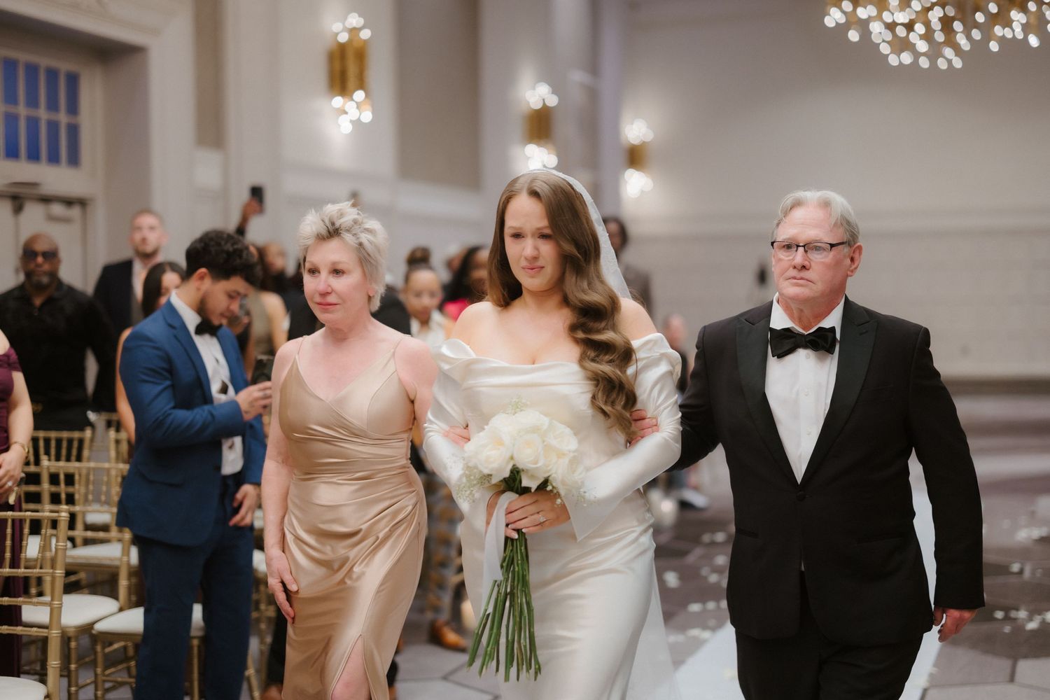 Bride in white gown walks down aisle with parents at elegant wedding ceremony.