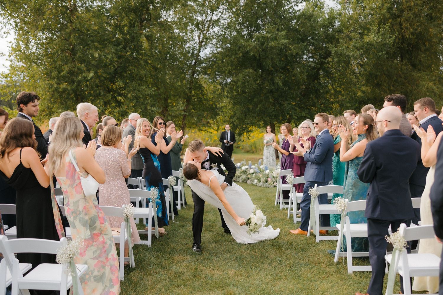 Groom dips bride for a kiss during wedding recessional while guests cheer and applaud outdoors.