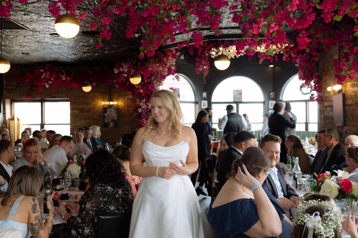 Smiling bride in strapless white gown addresses wedding guests beneath vibrant pink flower canopy indoors.