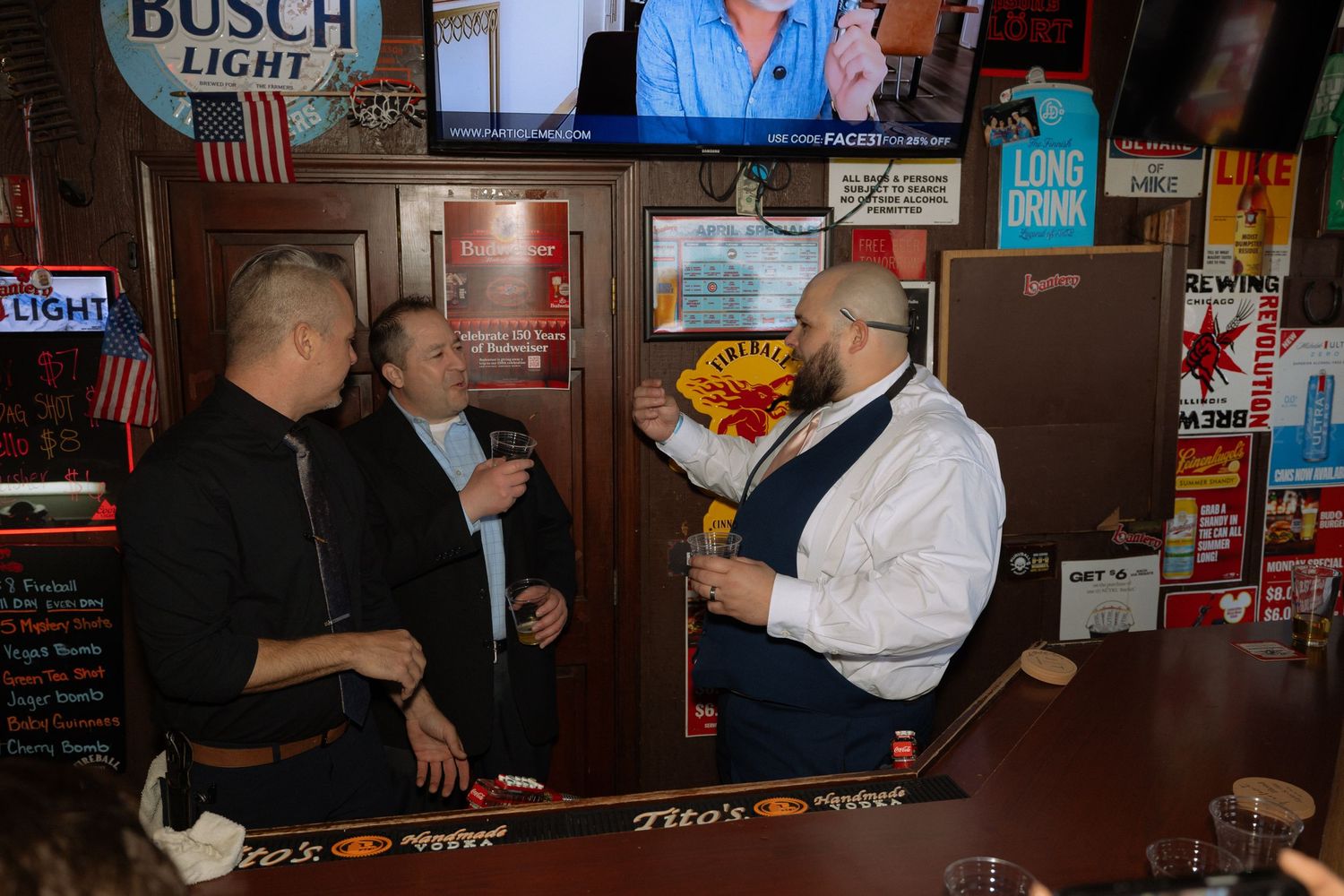 Three men conversing in a bar with Busch Light signs, wood paneling, and a Long Drink advertisement in the background.