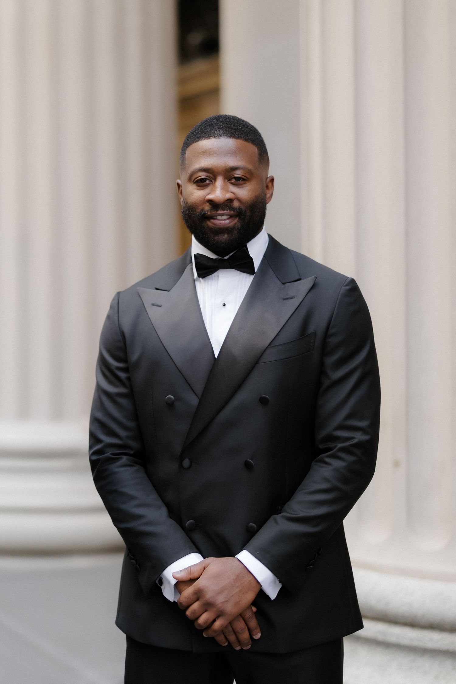 A man in a black tuxedo with bow tie smiles confidently in front of classical columns.