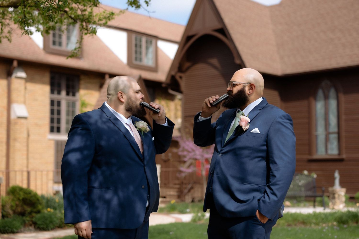 Two groomsmen in navy blue suits drinking outside a brick church on a sunny wedding day.