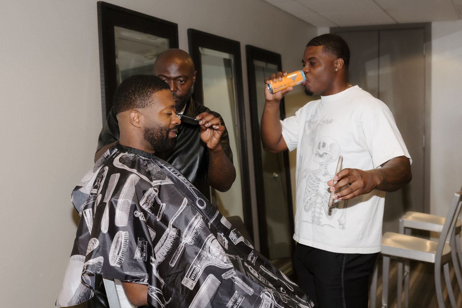 Three men in a barbershop, one getting a haircut while another drinks an orange beverage nearby.