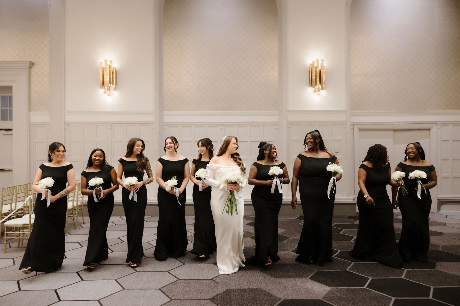 Bride in white gown with bridesmaids in black dresses holding white bouquets in elegant ballroom venue.
