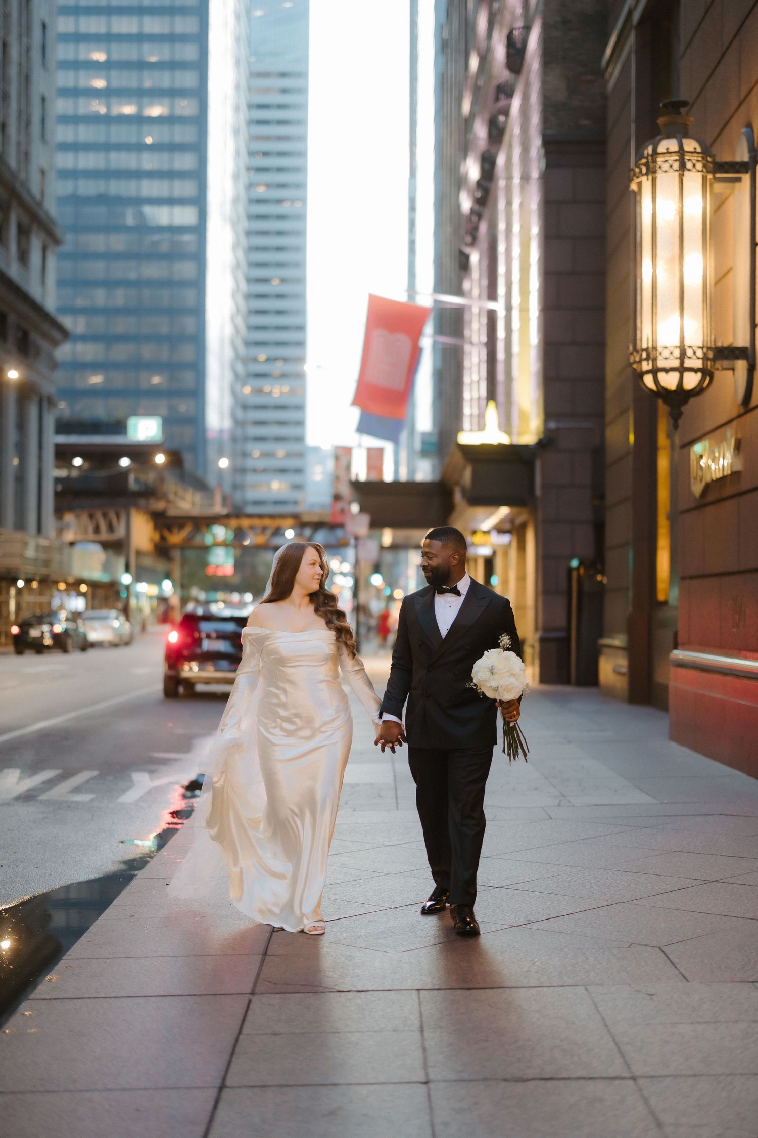 Bride and groom walk hand-in-hand on a city sidewalk at dusk, she in a flowing white gown, he in a classic black tuxedo.