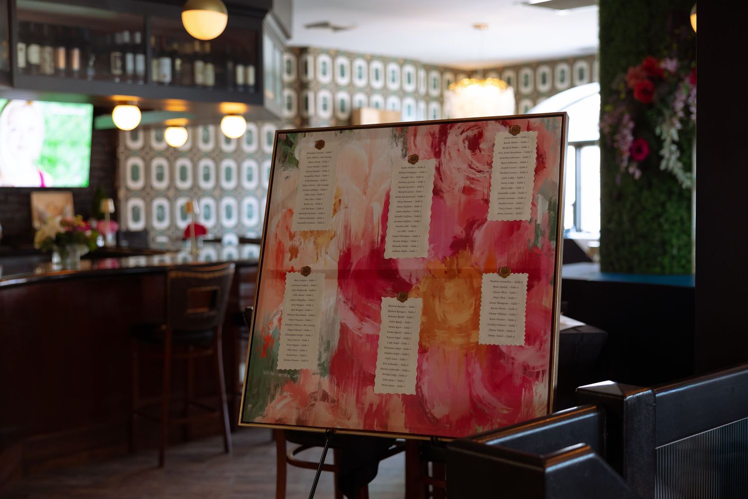 Floral seating chart display on easel in elegant bar venue with decorative wallpaper and warm lighting.