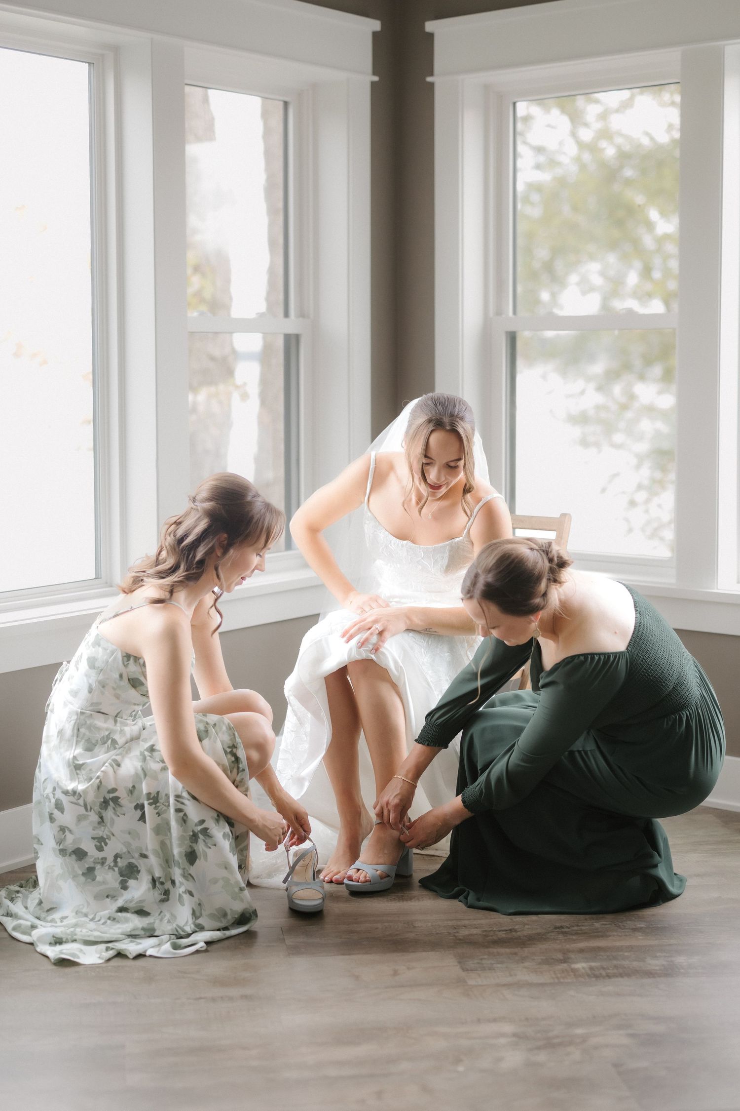 Two women kneel to help bride in white wedding dress and veil put on shoes by bright windows.