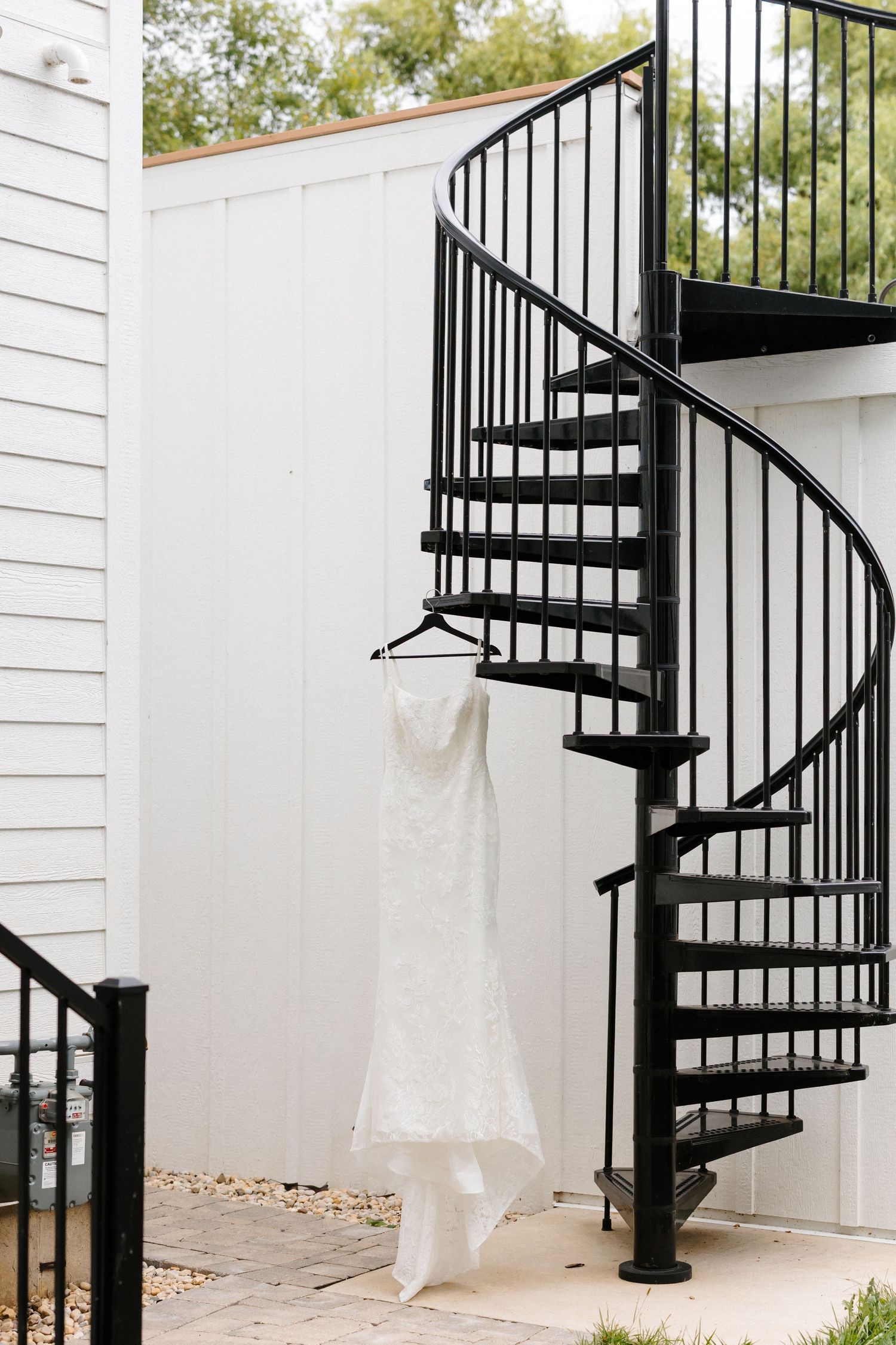 White lace wedding dress hanging from a black spiral staircase railing at an outdoor venue.