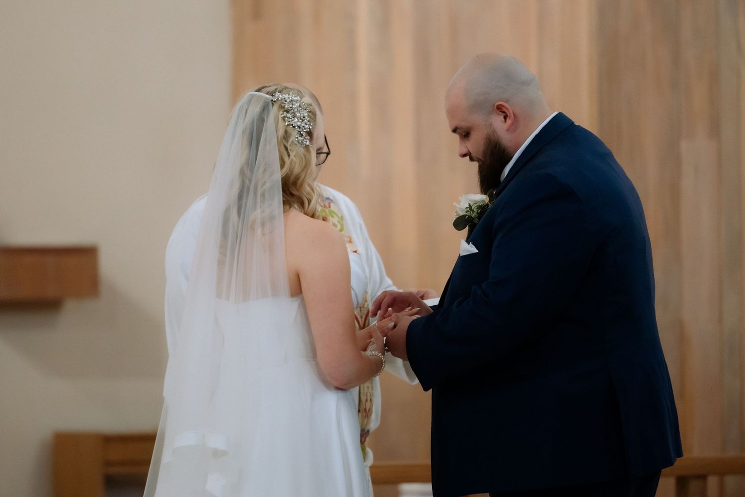 Bride and groom exchange rings at church altar, bride wearing white gown and veil, groom in navy suit.