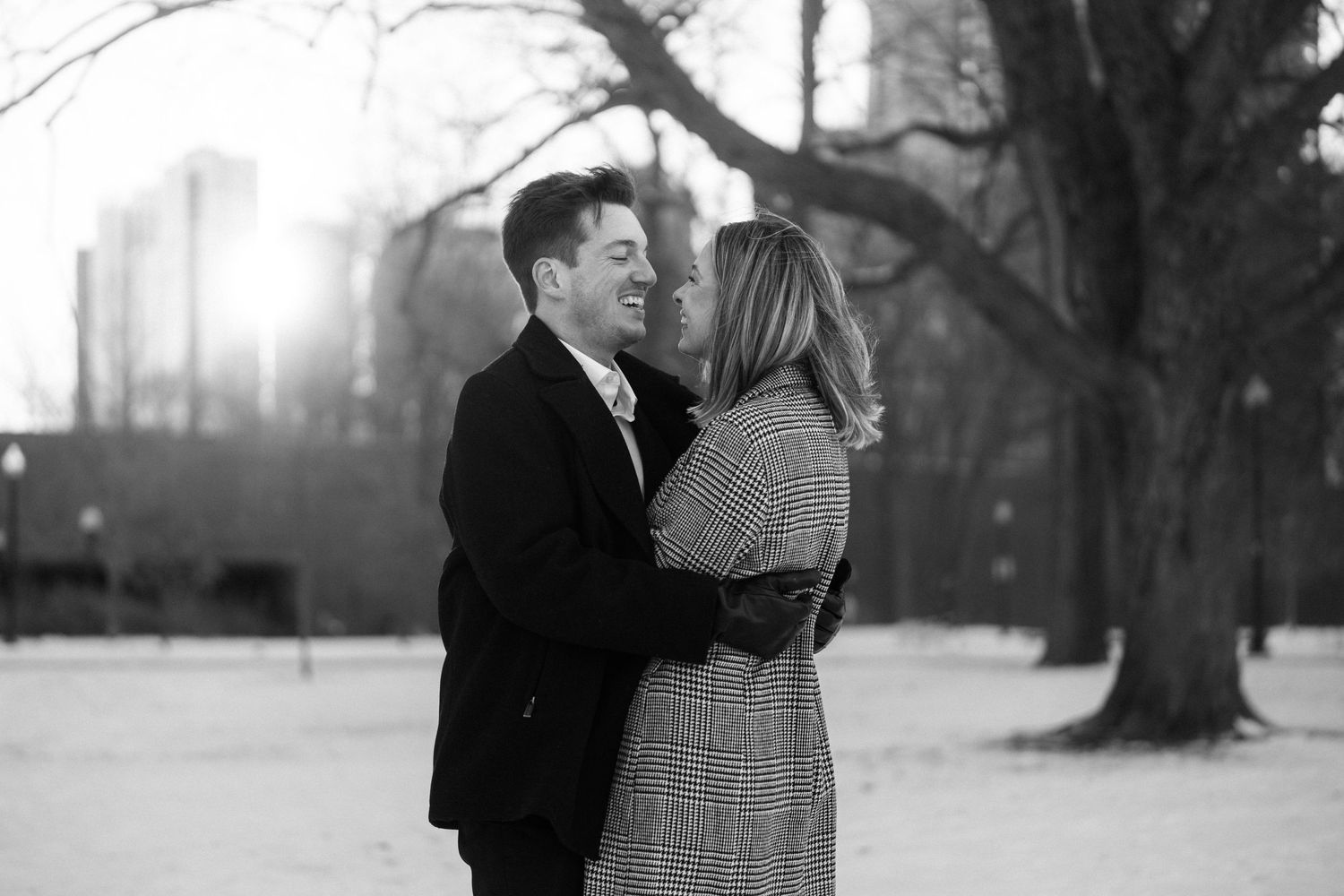 Black and white photo of couple about to kiss in snowy park with sunlight and city skyline behind them.