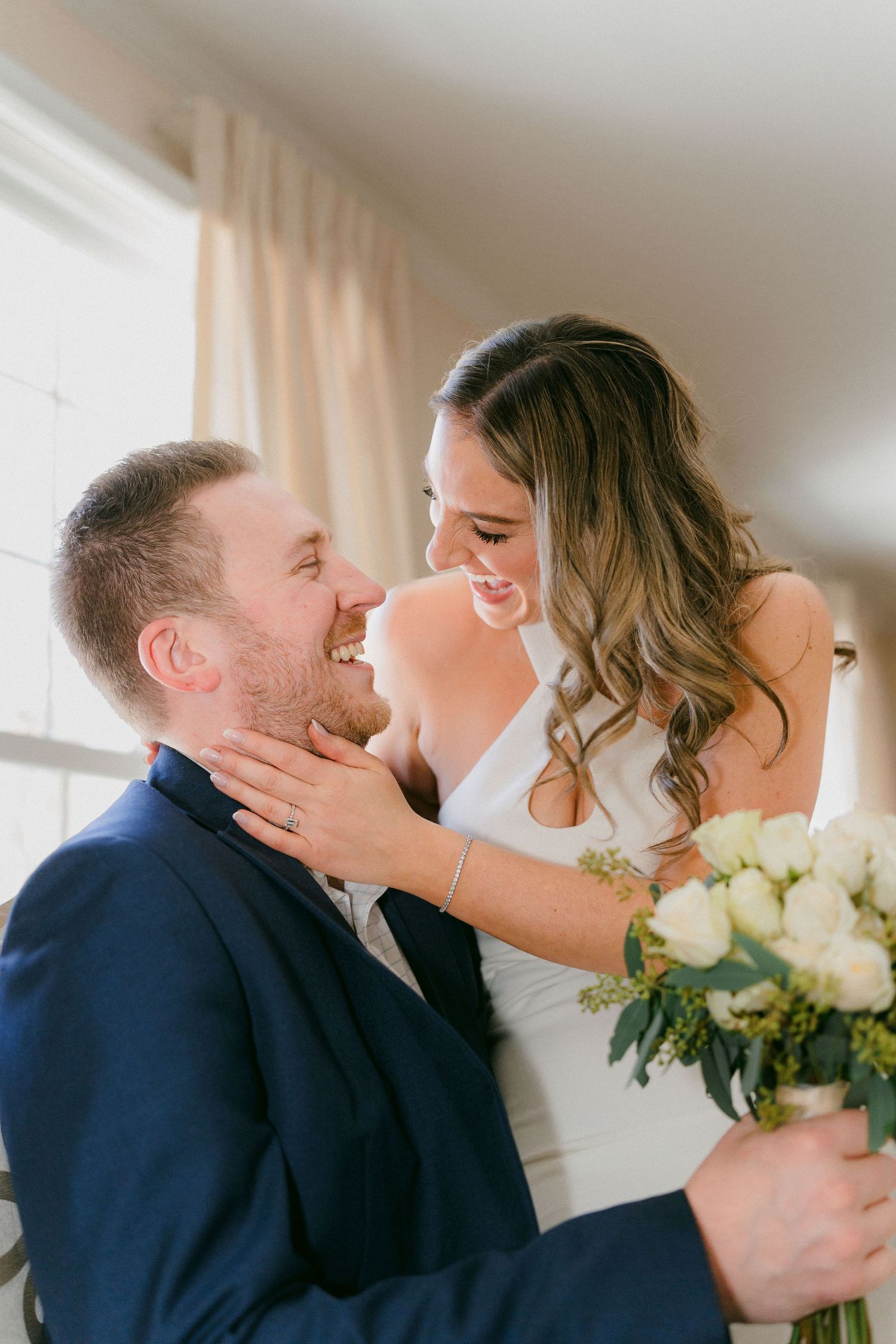 Laughing bride and groom share a joyful intimate moment with white rose bouquet by bright window.