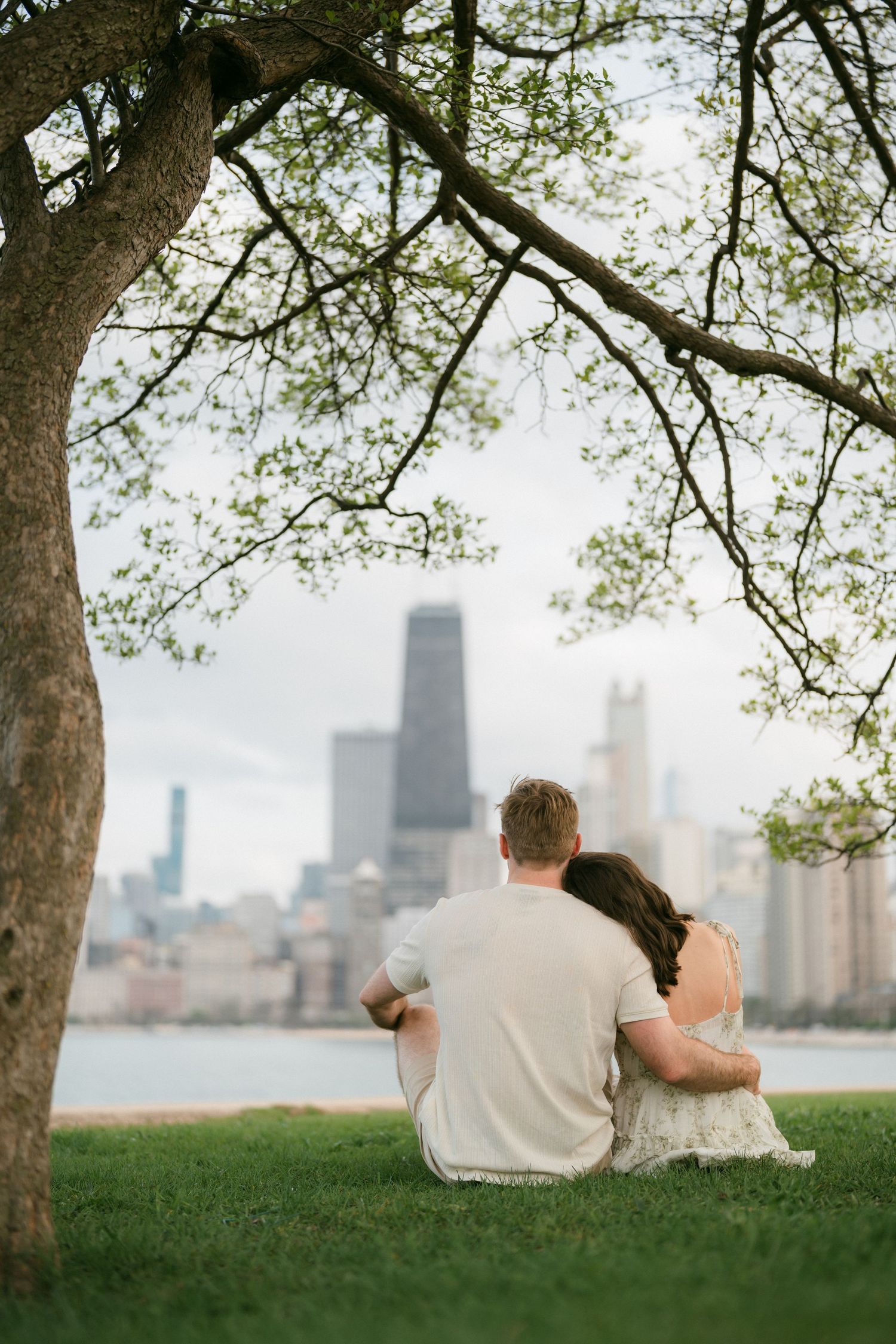 A couple sits embracing under a tree with a city skyline and lake in the background.