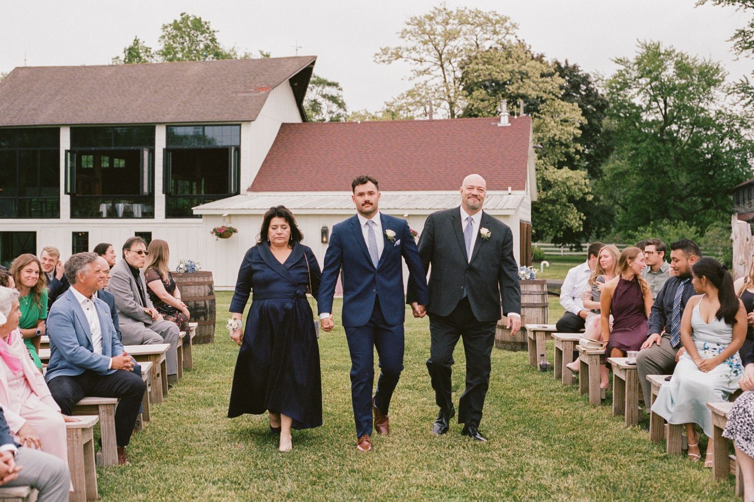 Groom walks down outdoor wedding aisle with parents, guests seated on wooden benches, barn backdrop.