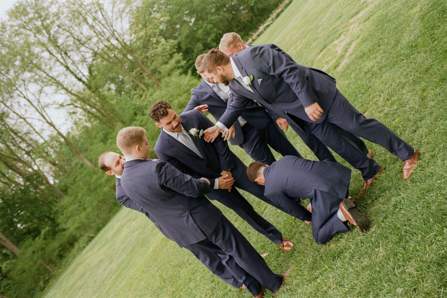 Groomsmen in navy suits celebrate outdoors on green grass with a fun group pose before a wedding.
